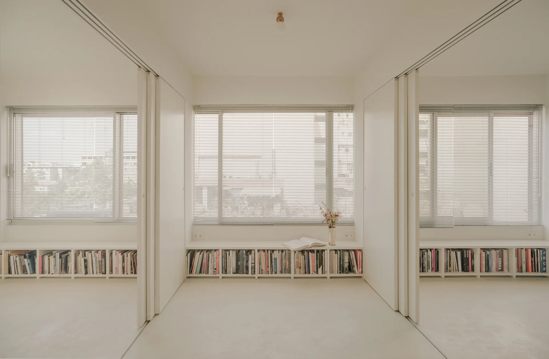 Three large windows with metal blinds overlook a minimalist white room featuring continuous low-profile bookshelves and sliding white partition panels.
