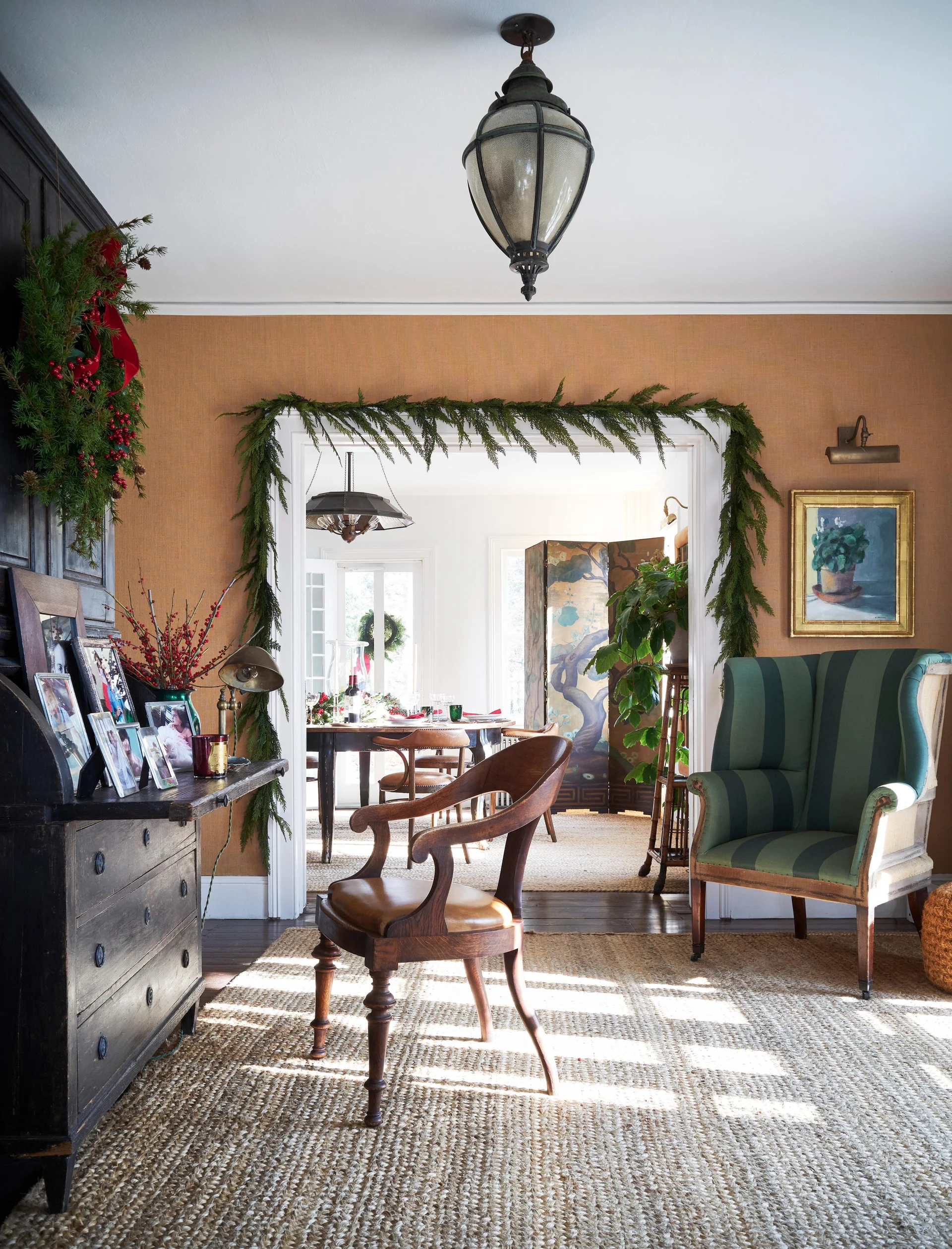 A green striped wingback armchair sits next to a doorway decorated with a natural pine garland, leading into a sunlit dining room.