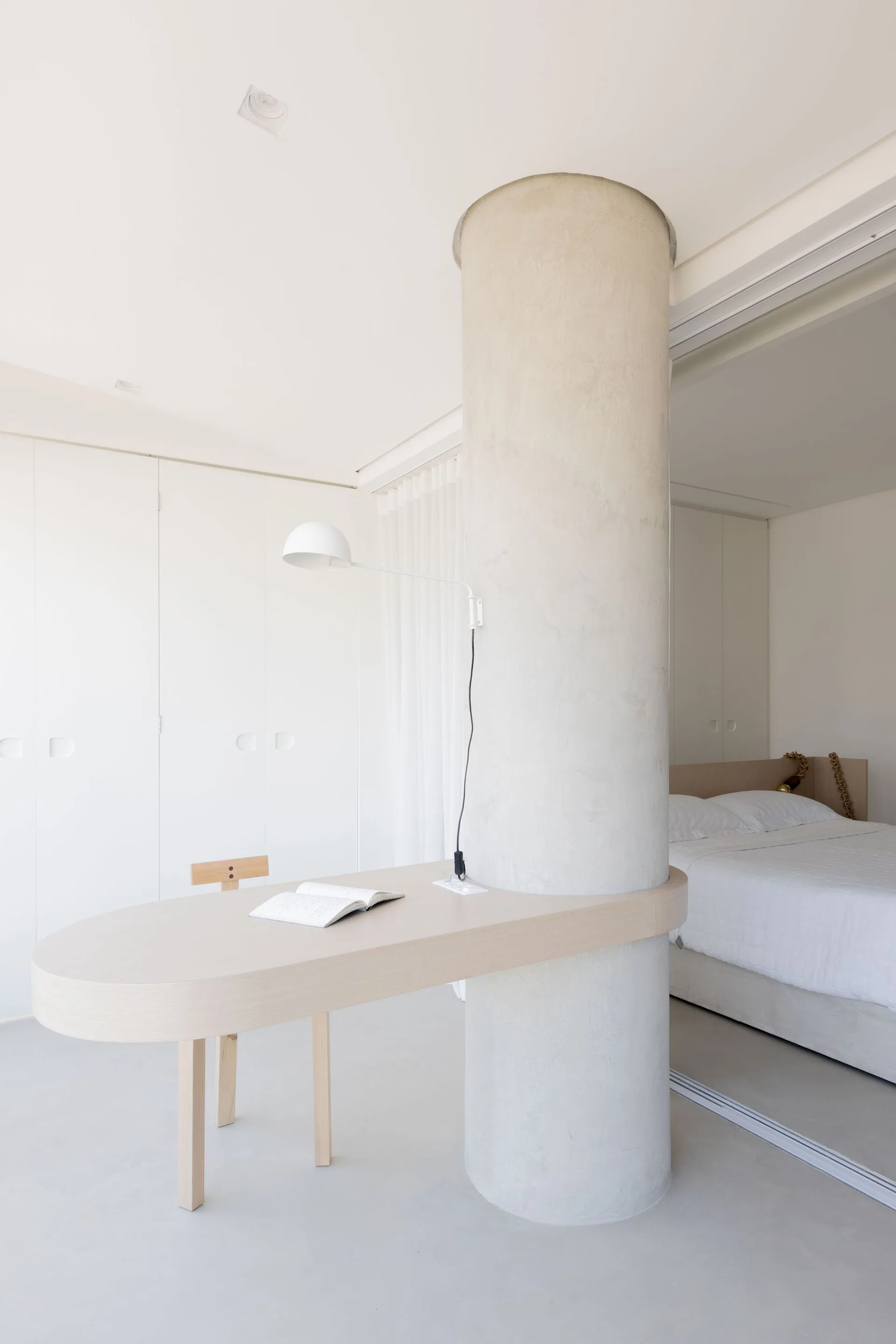 A light wood desk curves around a large round plaster column in a minimalist white bedroom featuring built-in wardrobes and a bed.