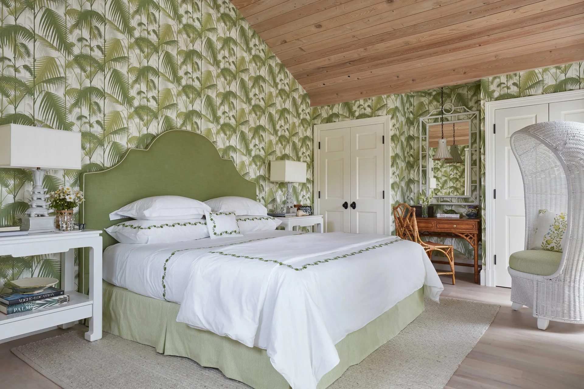 A bedroom features green palm-patterned wallpaper, an upholstered green headboard on a bed with white linens, and a slanted natural wood plank ceiling.
