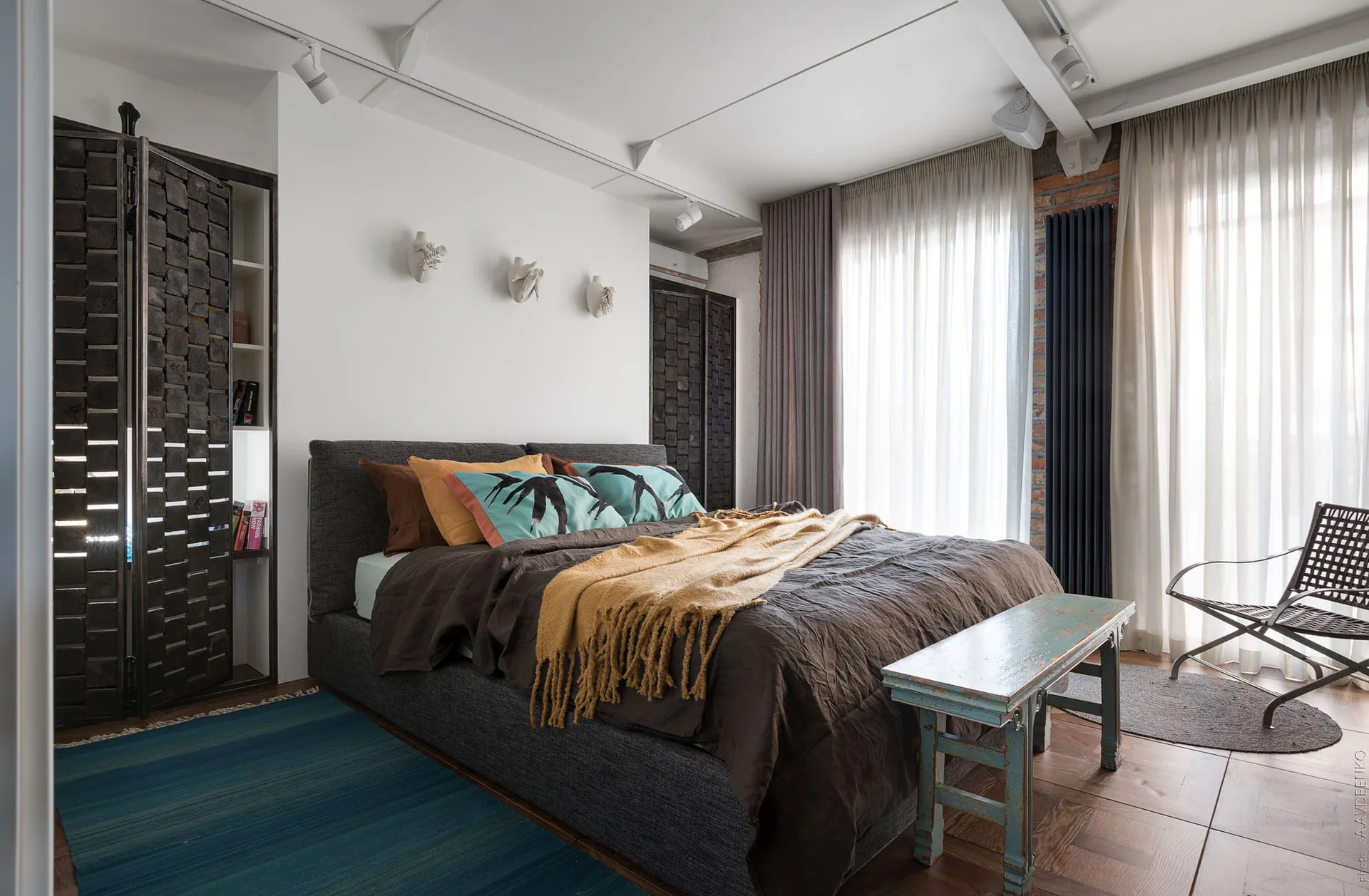 A bedroom featuring a dark upholstered bed with yellow and teal pillows, a distressed blue wooden bench, and exposed brick walls.