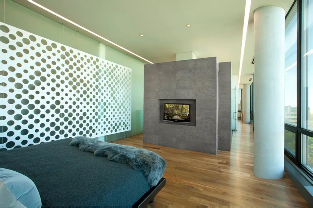 A modern bedroom features a dark bed, a gray stone block fireplace wall with a built-in television, and a white screen with circular cutouts.