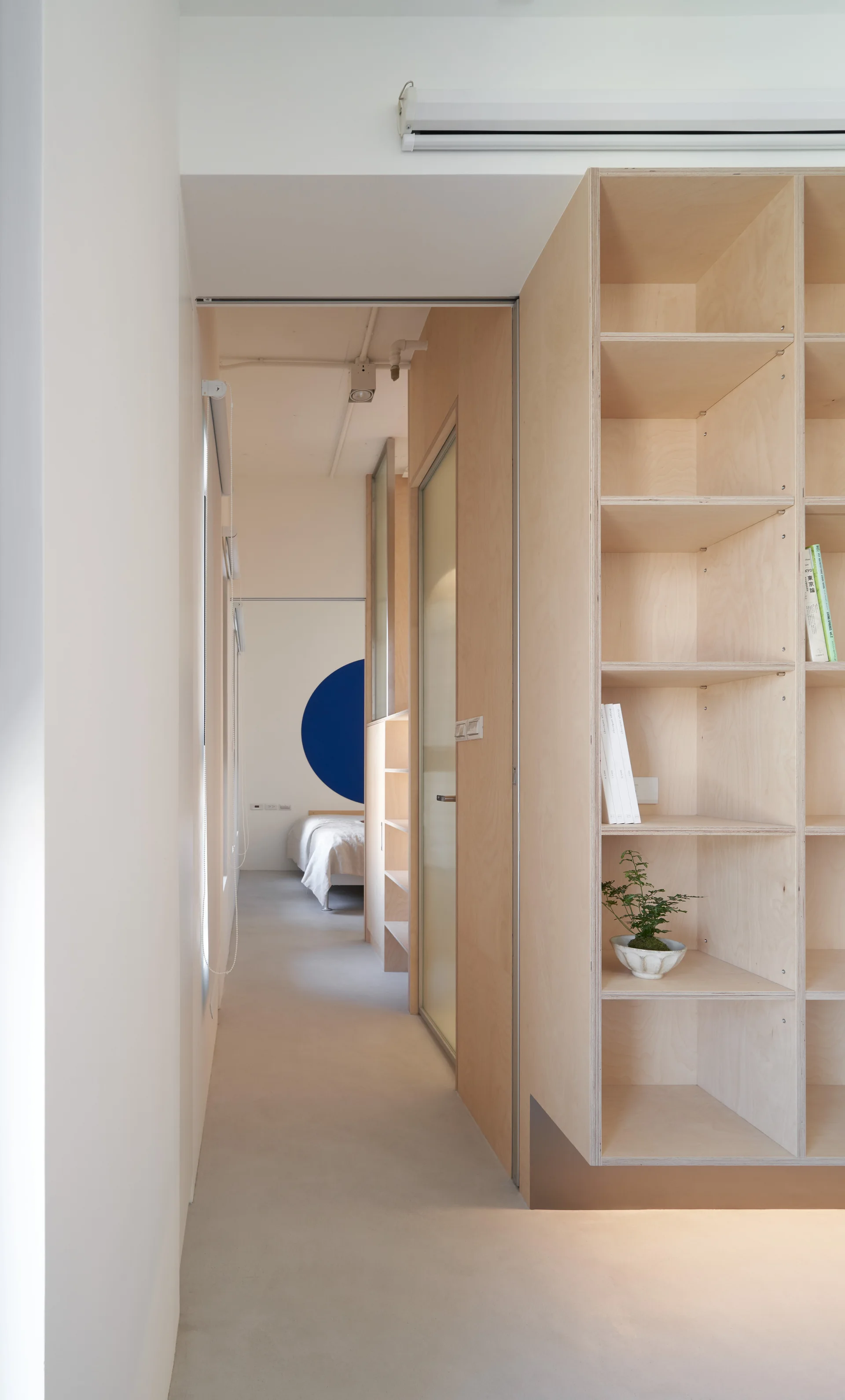 A minimalist white hallway features a tall light wood plywood shelving unit on the right, looking toward a bedroom with a blue circular wall graphic.