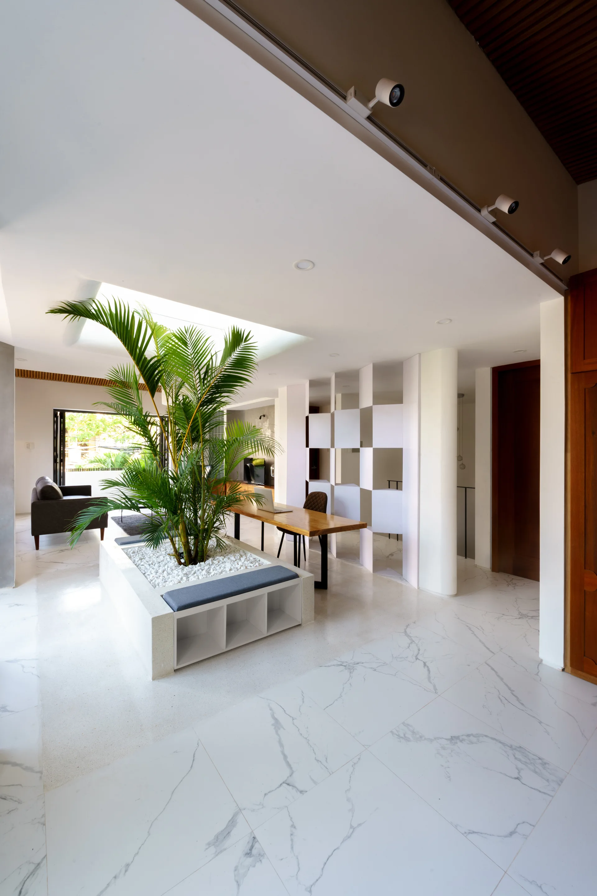 An open-plan room features a large built-in white planter with a tall green palm under a skylight, set on veined marble floors.