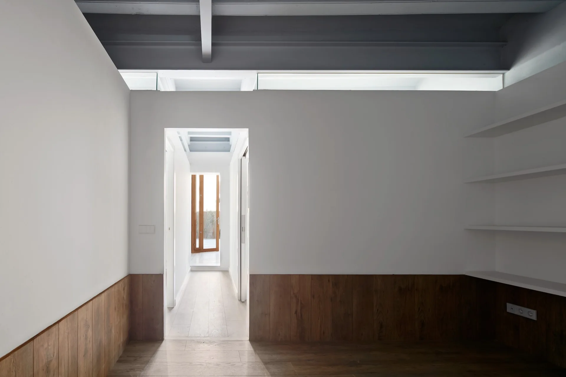 A minimalist hallway features white walls with dark wood wainscoting, white floating shelves, and exposed grey ceiling beams.