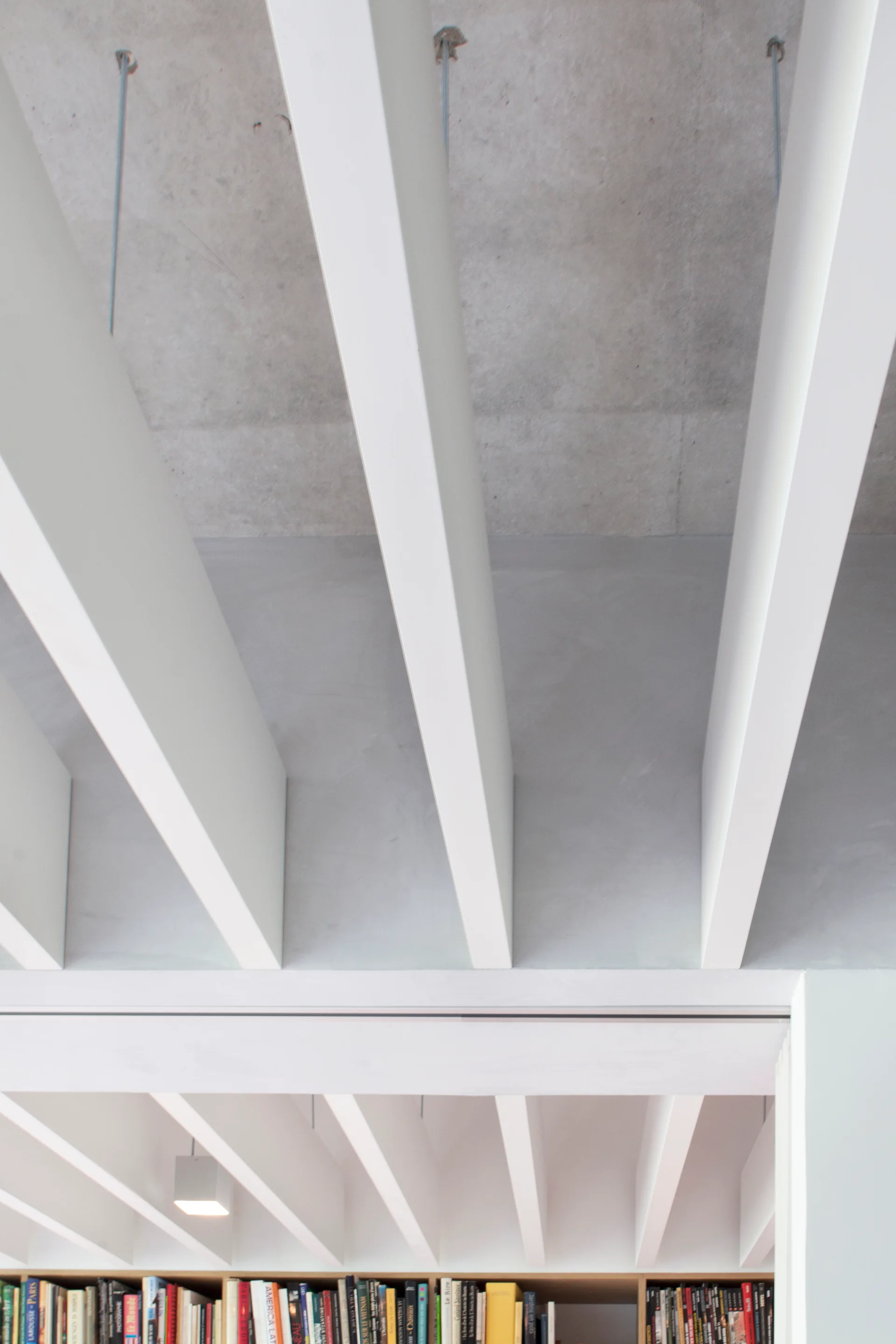 White painted ceiling beams are spaced evenly across a raw grey concrete ceiling above a modern built-in bookshelf filled with books.