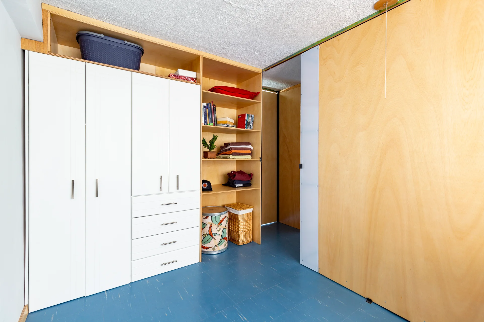 A closet area featuring built-in white wardrobe doors, integrated plywood shelving, a plywood sliding door, and a matte blue floor.