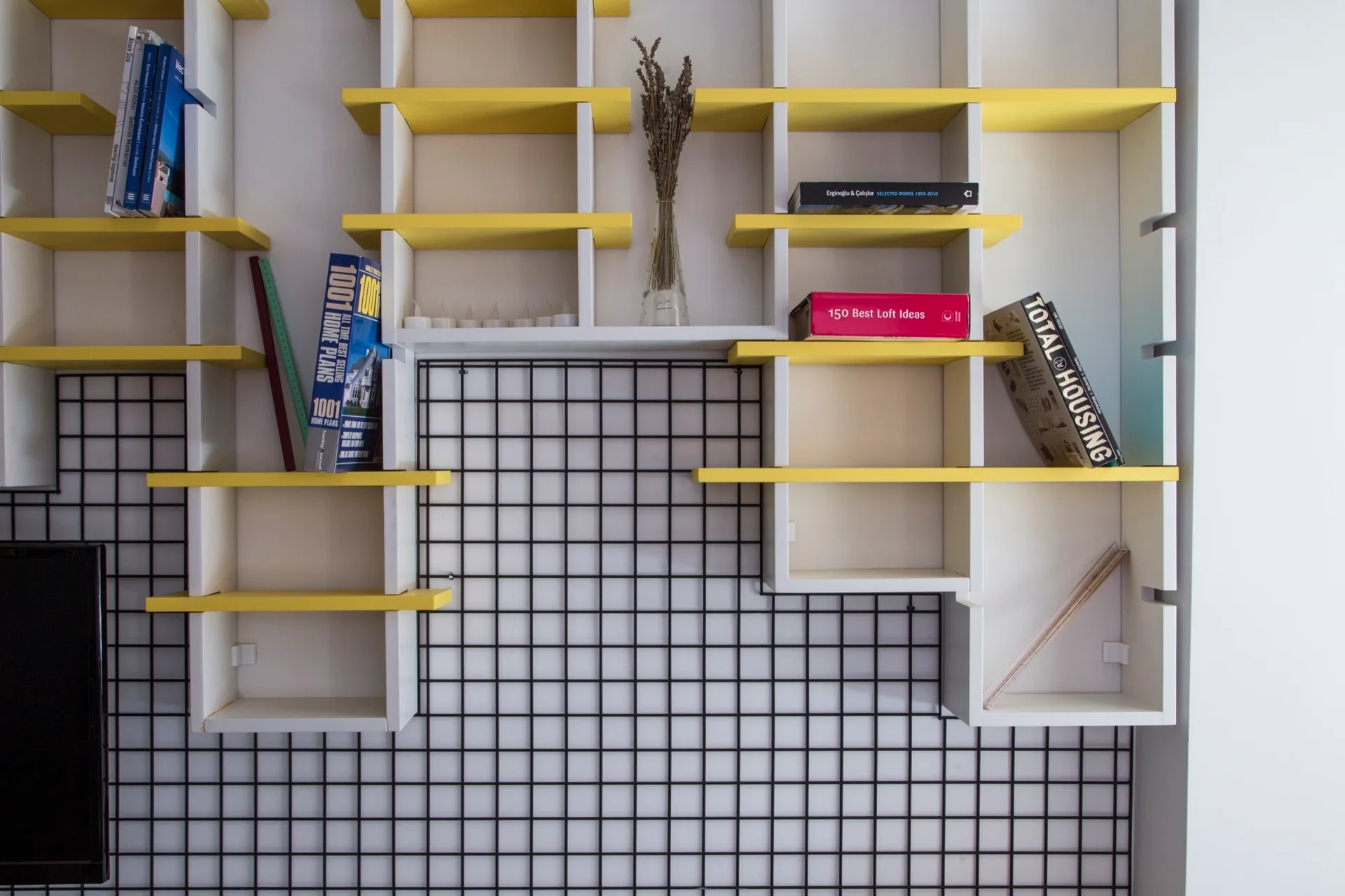 A modular white wall shelving unit with bright yellow shelves and a black wire grid background holds books and a vase.