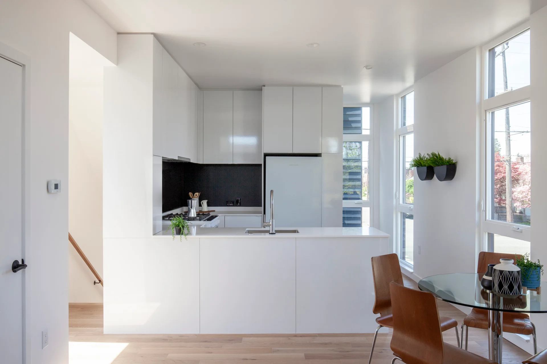 Minimalist white kitchen with glossy cabinets, a black backsplash, a white island, and a glass-top dining table with bentwood chairs.