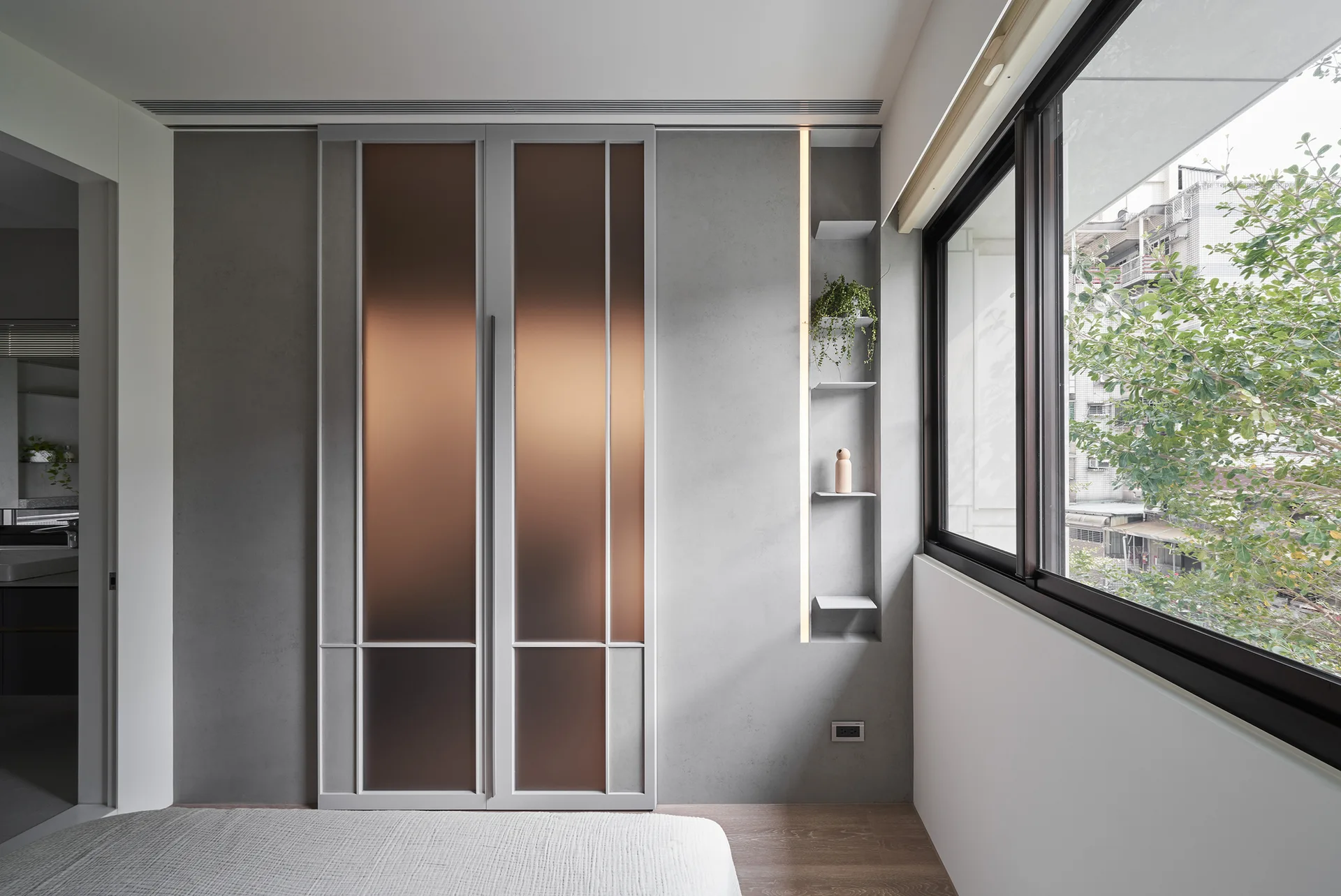 A minimalist bedroom featuring gray wardrobe doors with amber glass panels, a recessed shelving unit, and a window.