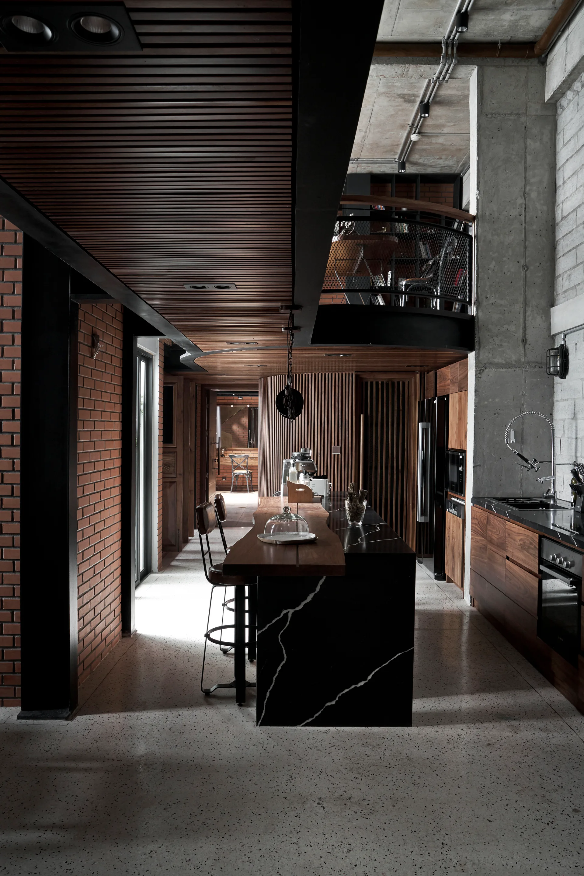 An industrial kitchen features a black veined marble island with bar stools, exposed red brick walls, concrete pillars, and a dark wood slatted ceiling.