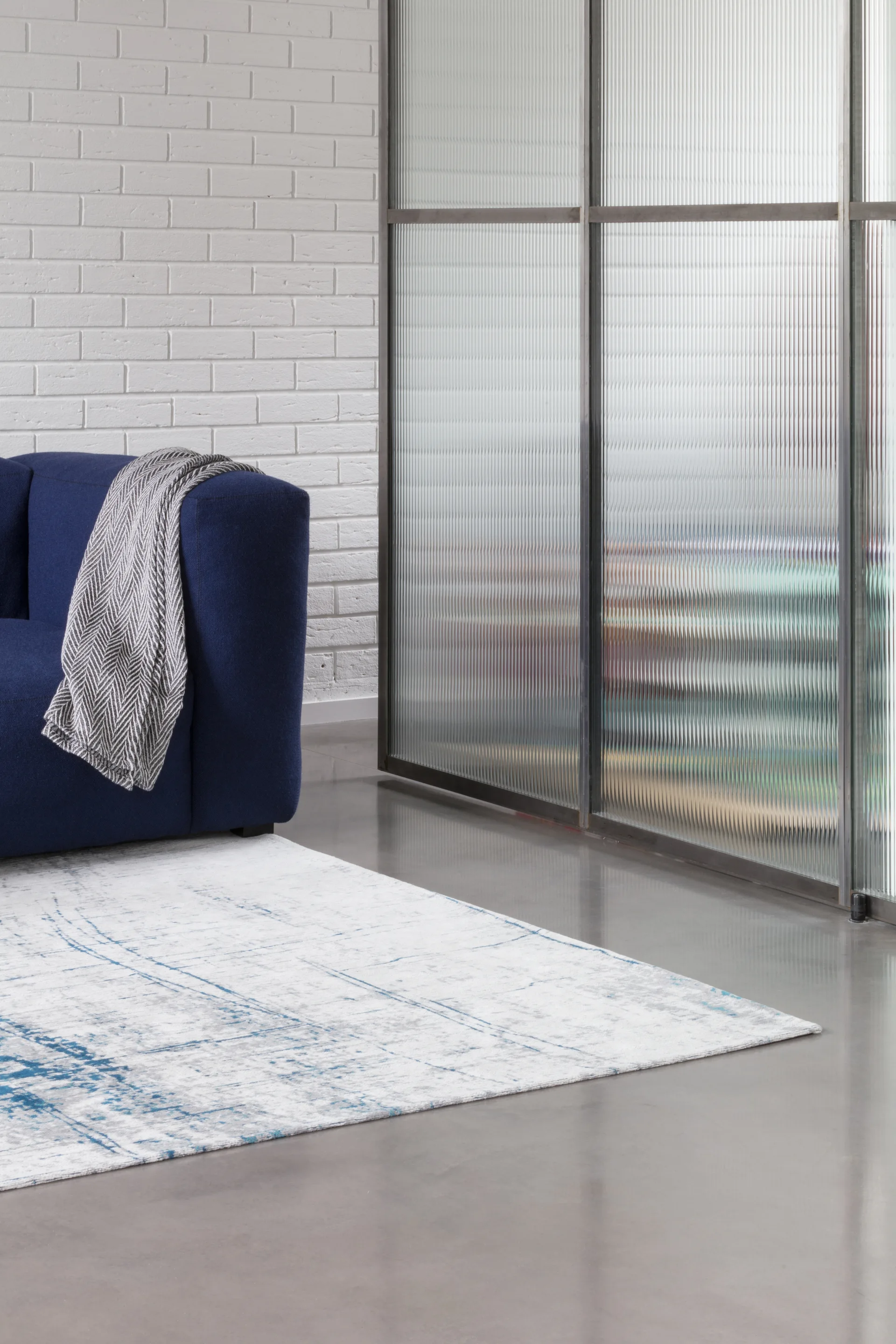 A blue sofa with a chevron throw blanket sits next to a metal-framed ribbed glass partition on a polished concrete floor.