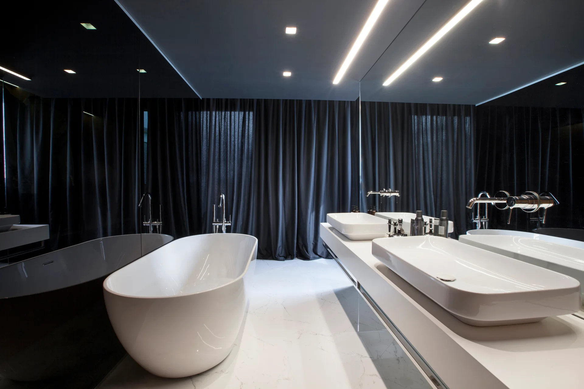 A sleek modern bathroom features a white freestanding bathtub, a floating vanity with double sinks, and dark floor-to-ceiling curtains.