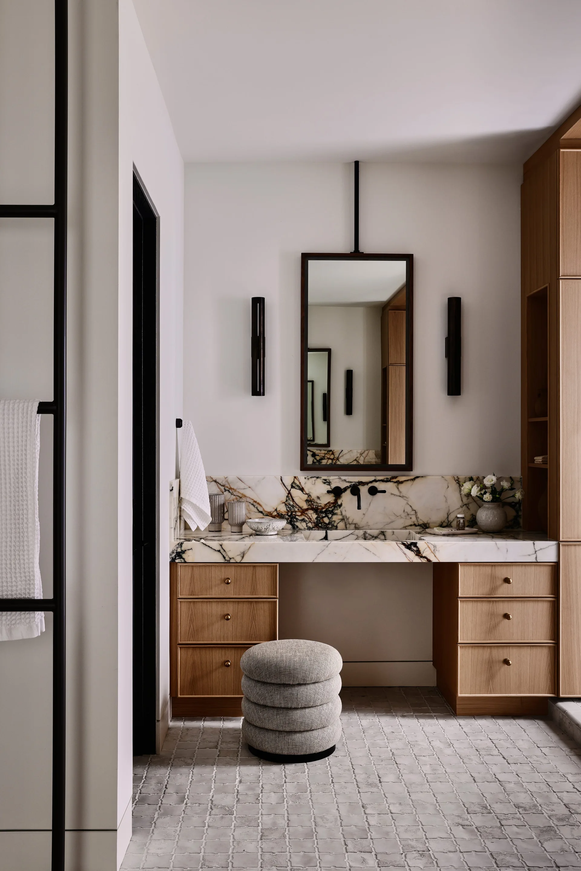 A modern bathroom vanity with light oak cabinetry, a heavily veined marble countertop and backsplash, a rectangular mirror, and a grey fabric stool.