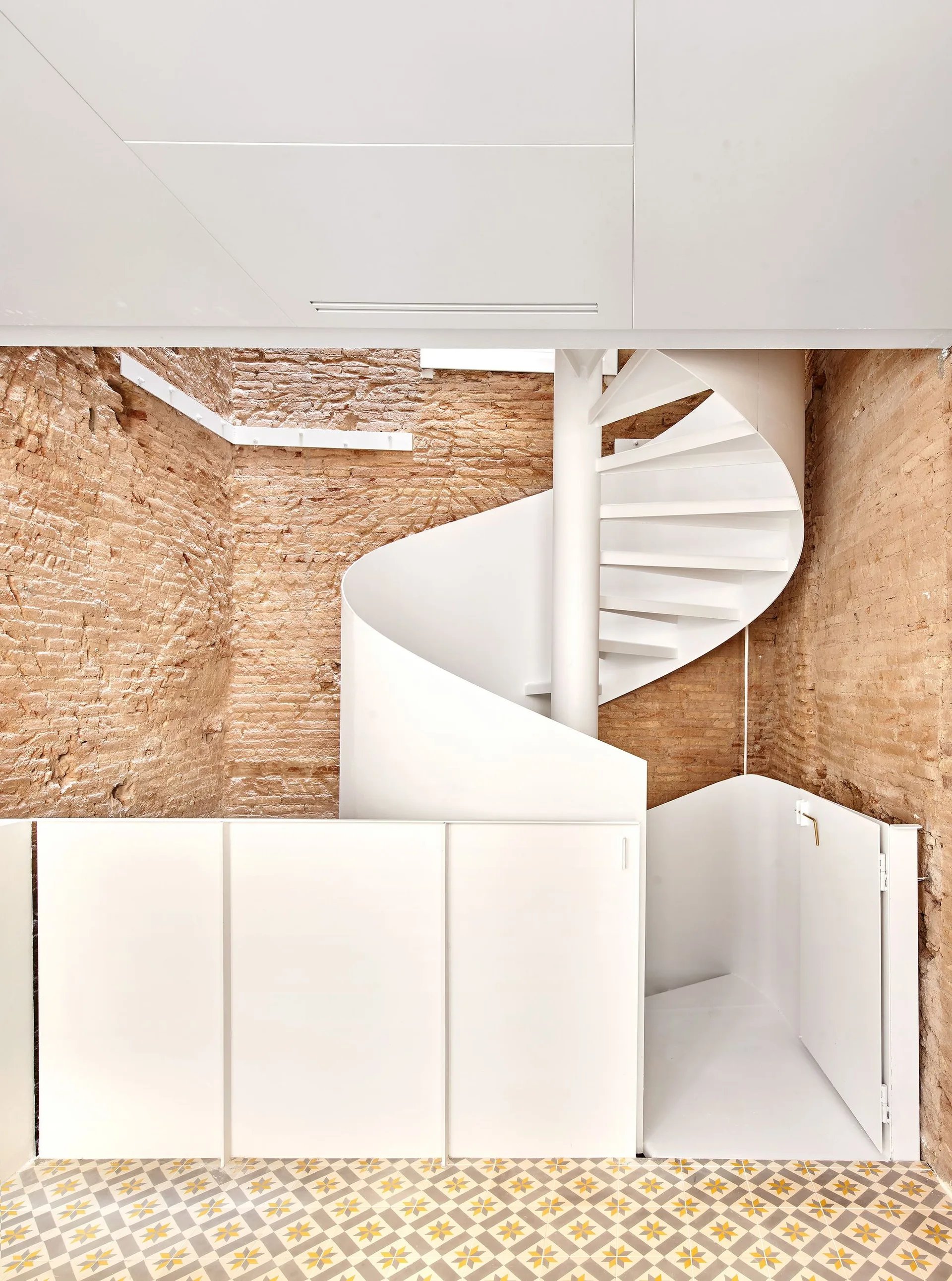 A modern white spiral staircase stands next to smooth white cabinets against a rustic exposed brick wall and geometric tiled flooring.