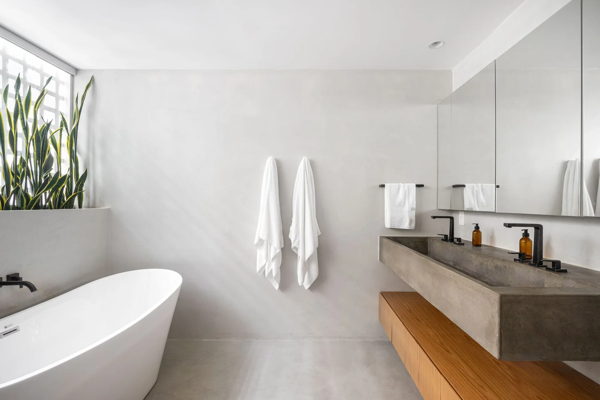 A contemporary bathroom features a white freestanding bathtub, a concrete trough sink on a floating wood cabinet, and smooth grey plaster walls.