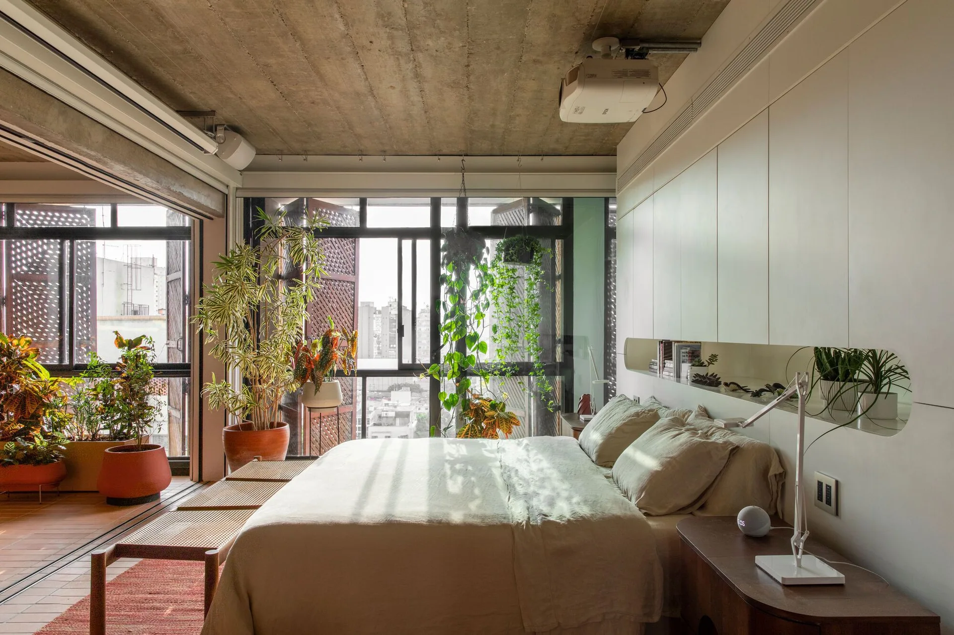 A modern bedroom with an exposed concrete ceiling, beige linen bedding, a white built-in storage wall, and a balcony filled with potted plants.