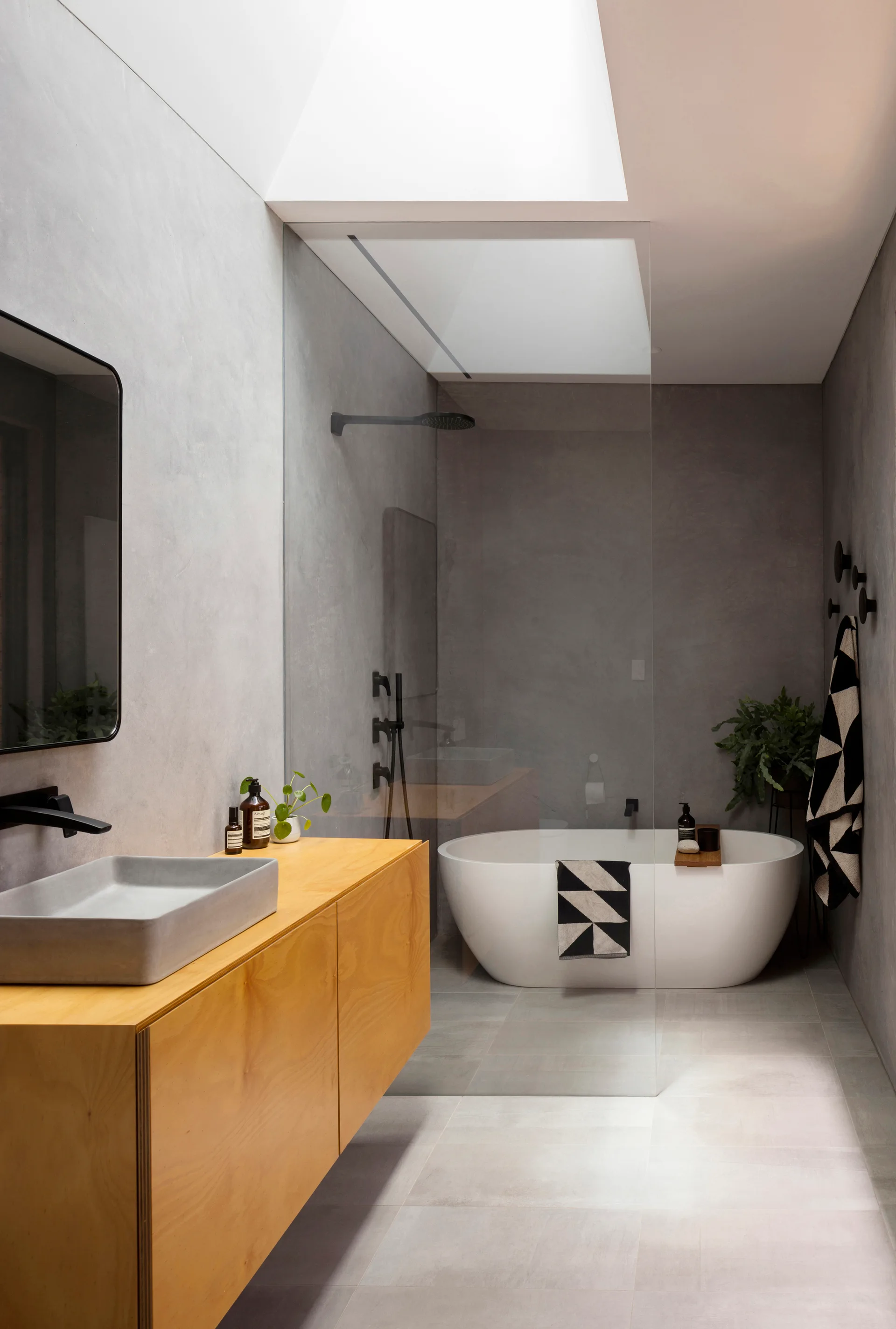 A modern bathroom features a floating light wood vanity with a grey basin sink, a freestanding white tub under a skylight, and concrete-finish walls.