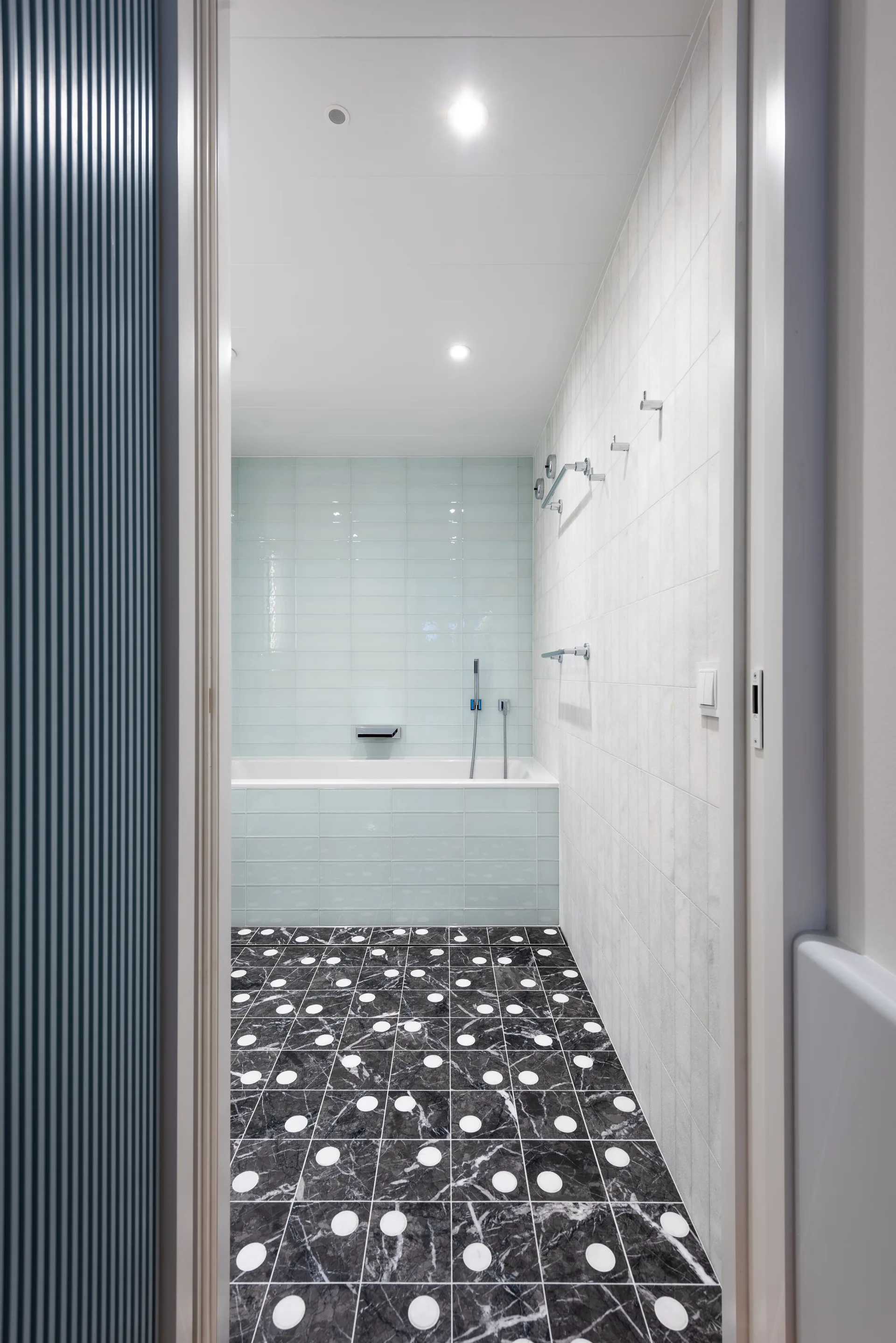 A modern bathroom with a white bathtub surrounded by glossy pale blue wall tiles and a black marble floor featuring white polka dots.