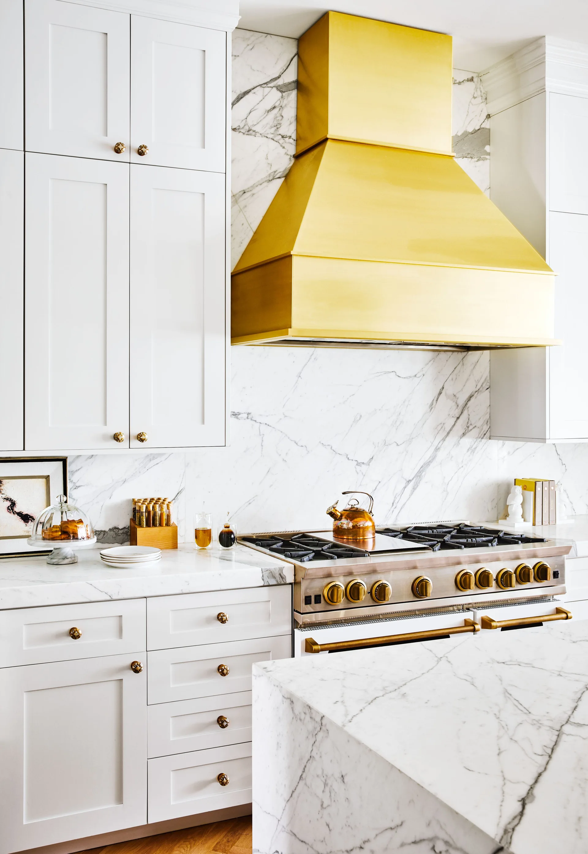 A kitchen featuring white shaker cabinets, a prominent gold range hood, a marble slab backsplash, and a professional gas stove.