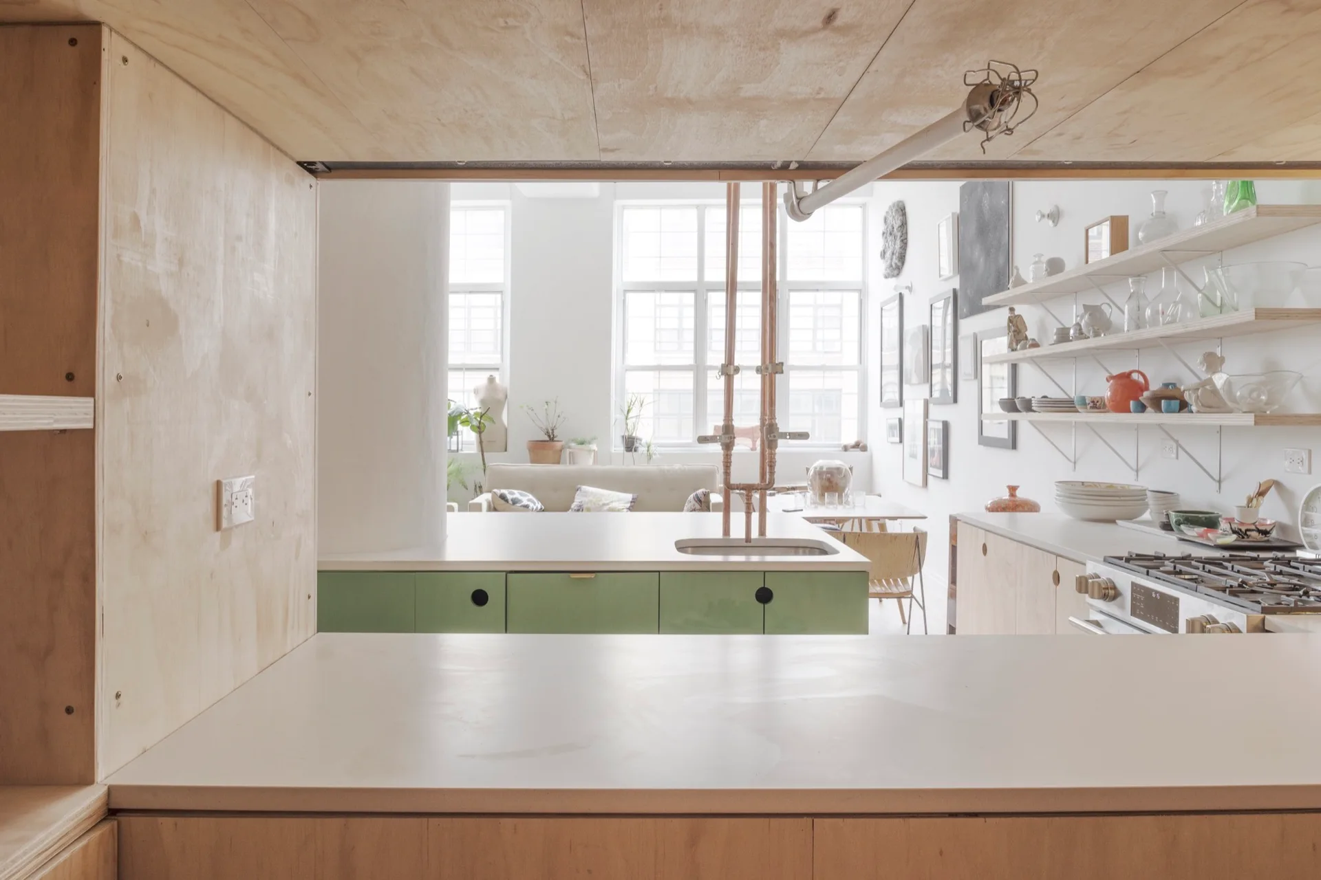 A bright kitchen featuring plywood ceiling panels, a green island with copper pipes, and a view of a sunlit living space.