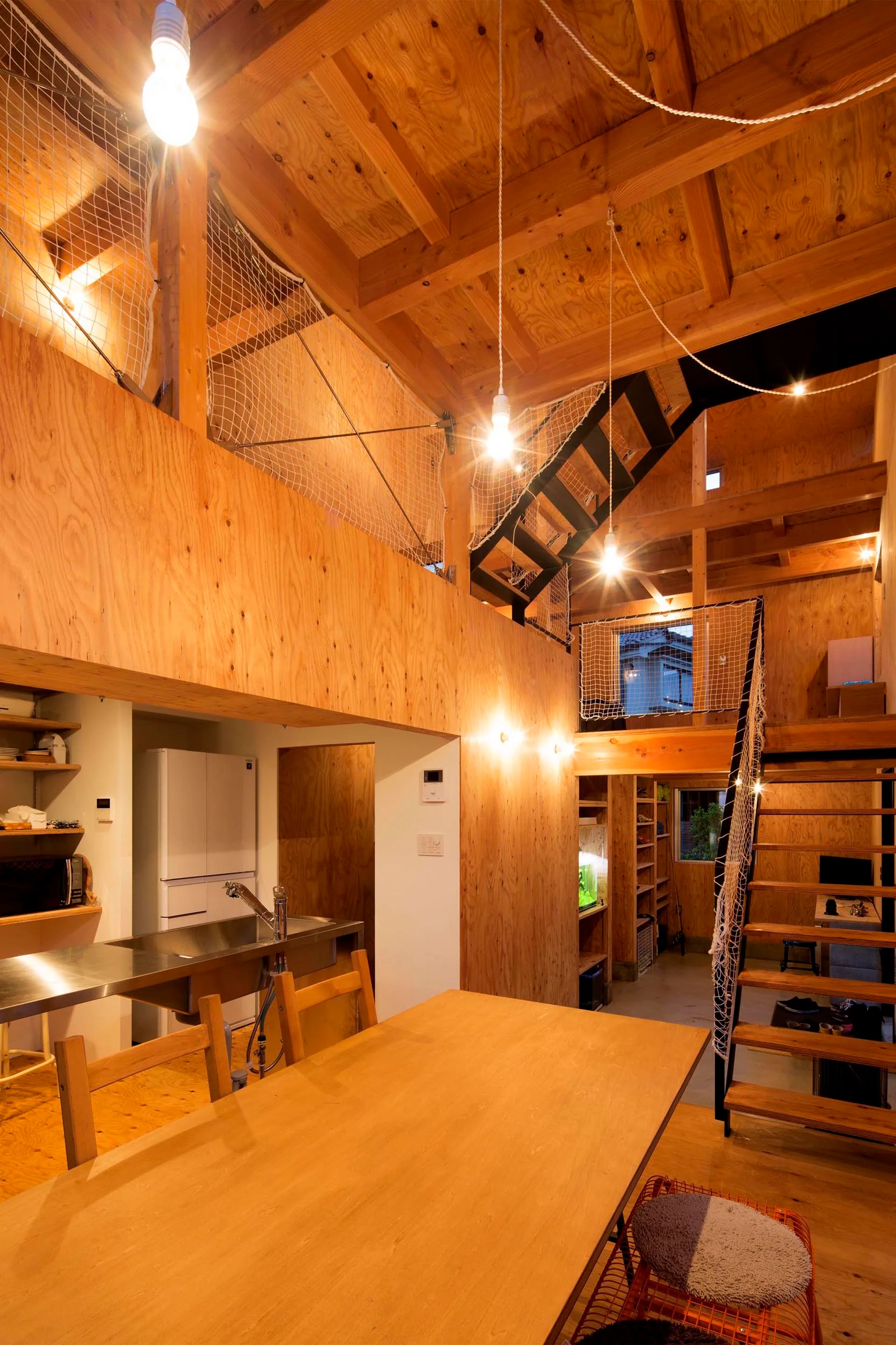A modern loft interior featuring plywood walls, a dining table, a stainless steel kitchen counter, and a mezzanine with safety netting.