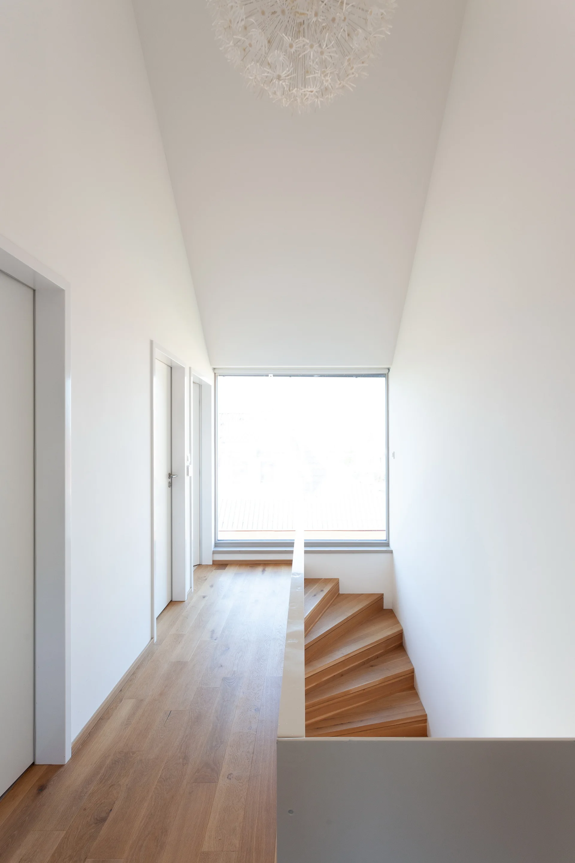 A bright minimalist hallway with white walls and light wood floors features a wooden staircase, a large window, and a white sculptural chandelier.