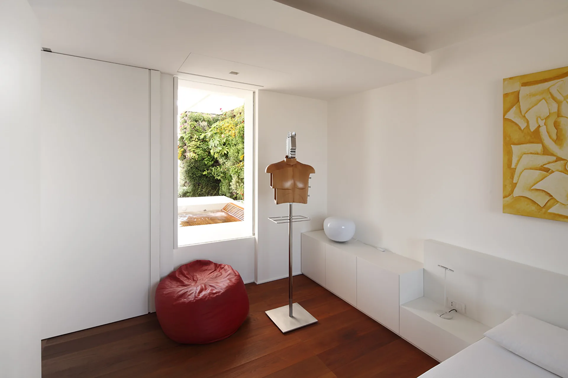 A minimalist bedroom with dark wood floors, low-profile white storage cabinets, a red bean bag chair, and a narrow window looking out to a garden.