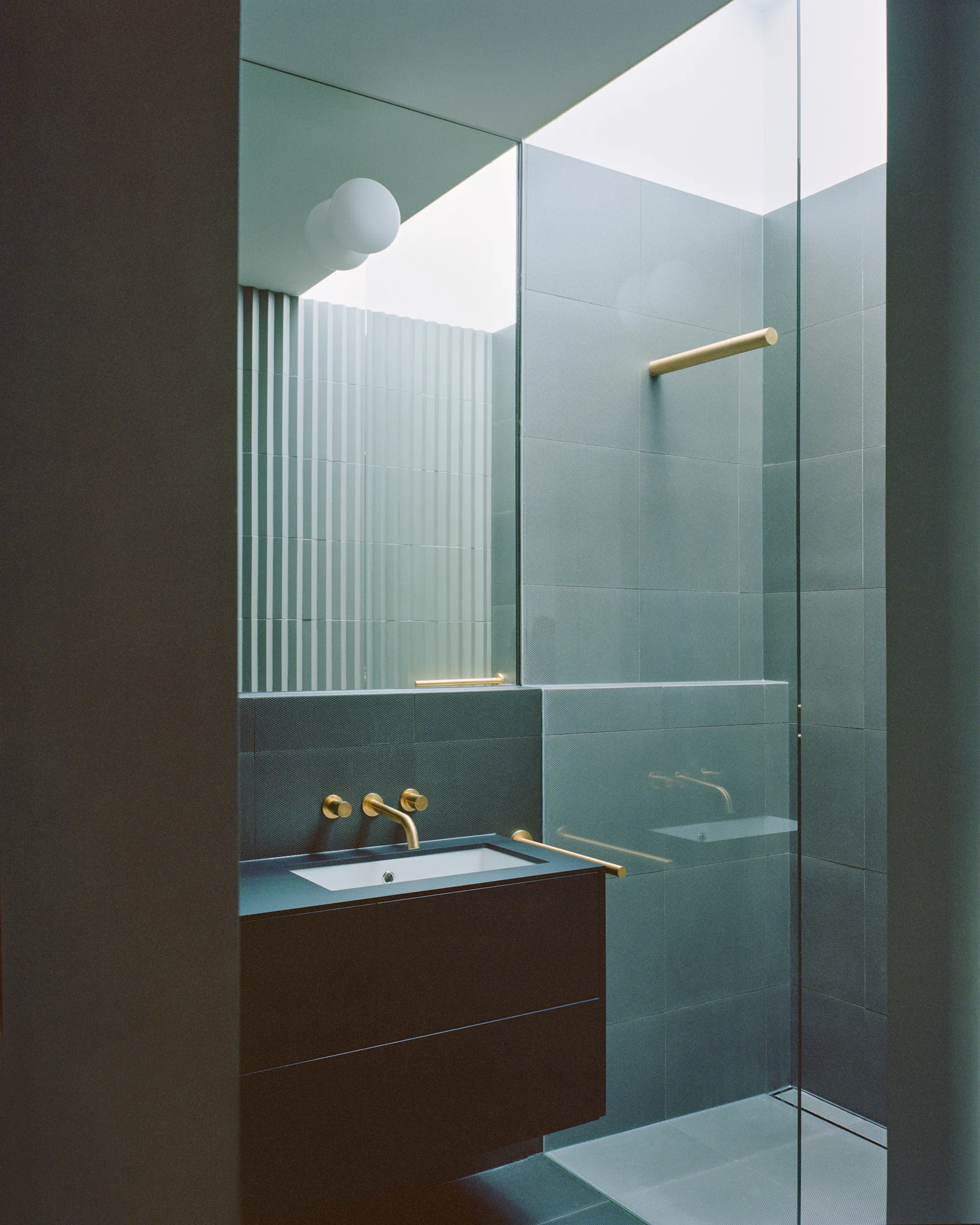 A contemporary dark gray bathroom features a wall-mounted vanity with brass fixtures, a glass shower partition, and tiled walls under a skylight.