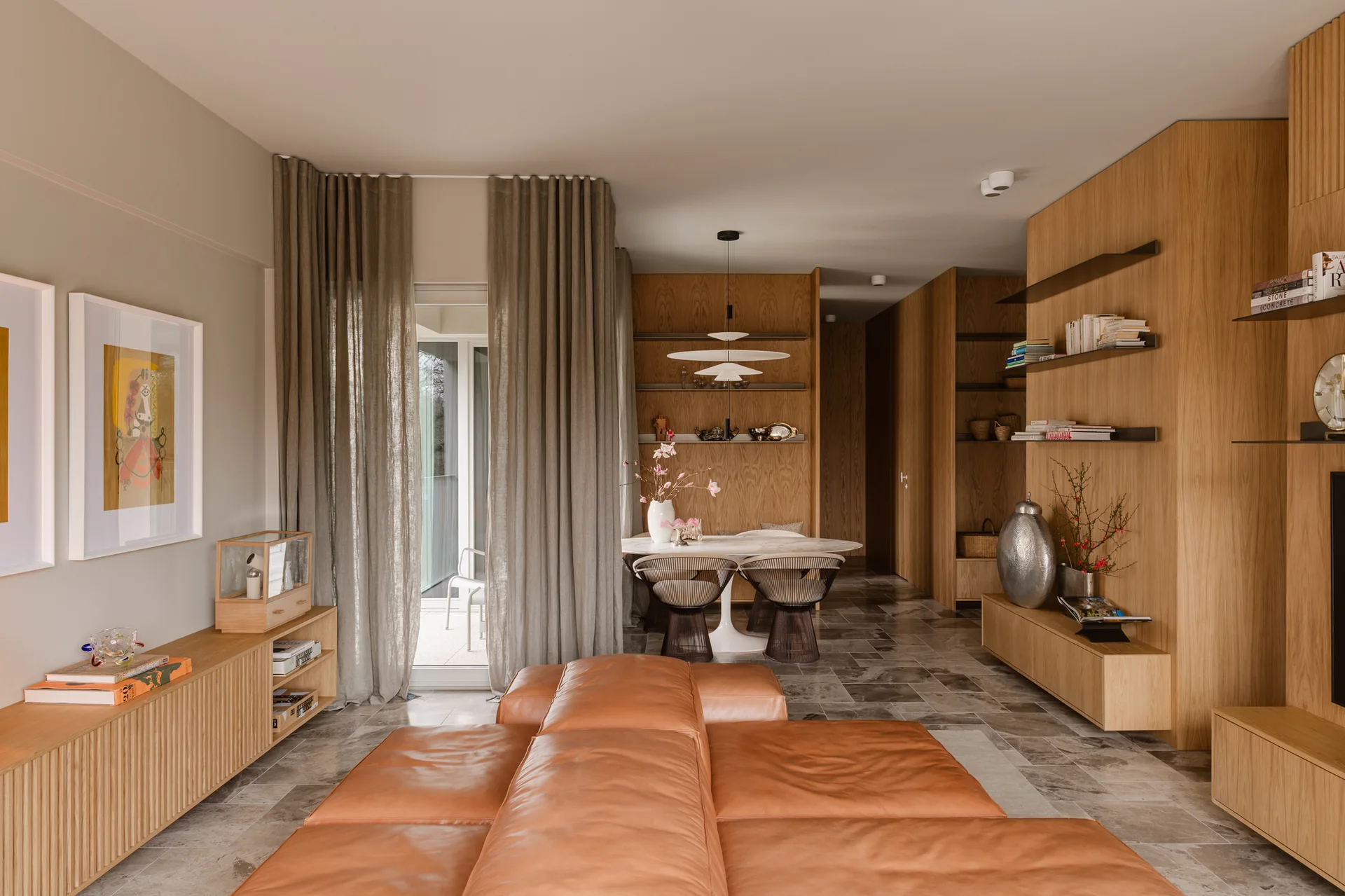 A modern living room features a modular tan leather sofa, stone tile flooring, light wood wall paneling, and a dining nook in the background.