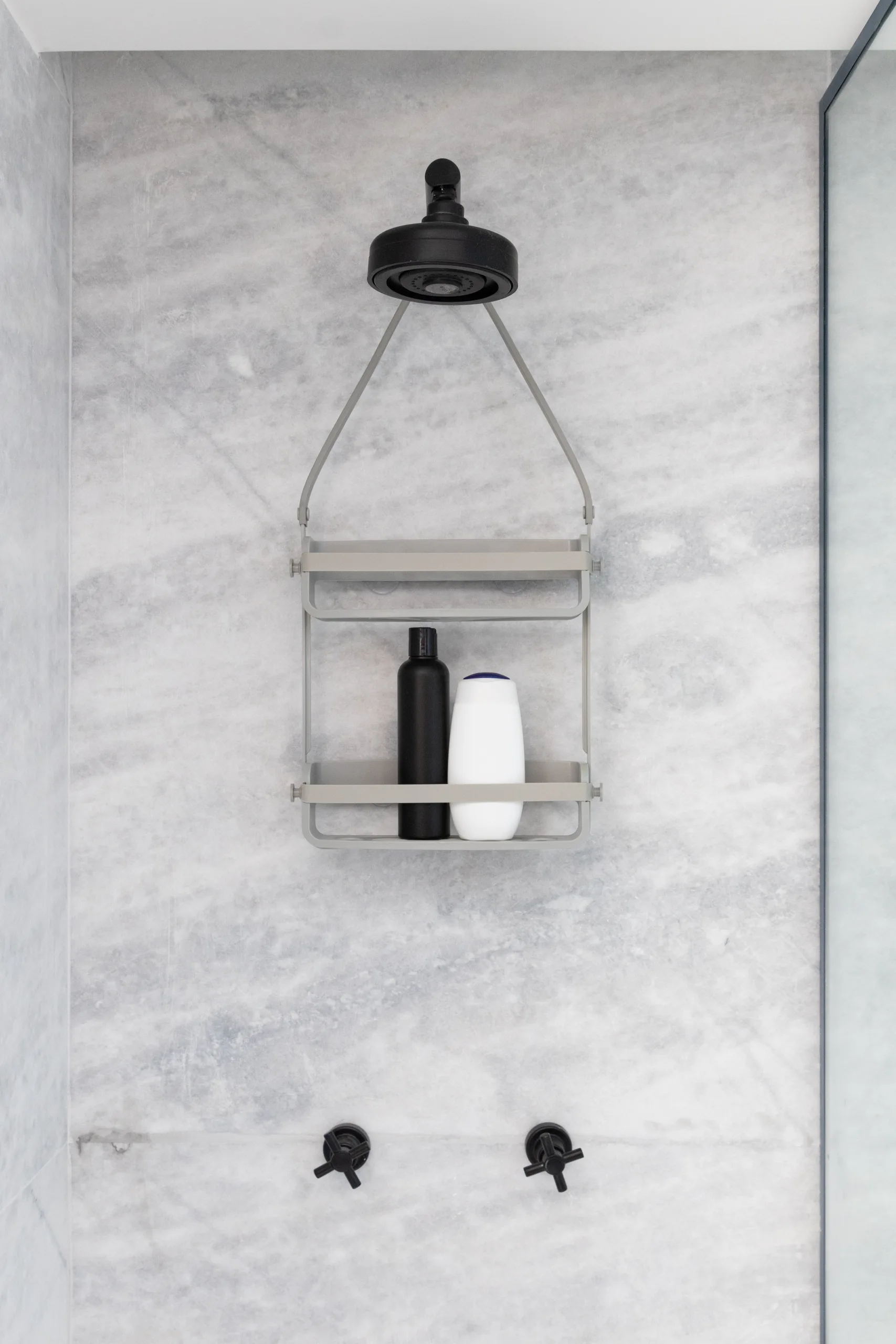 A grey hanging caddy holds bottles against a veined grey marble shower wall with a matte black shower head and matching faucet handles.