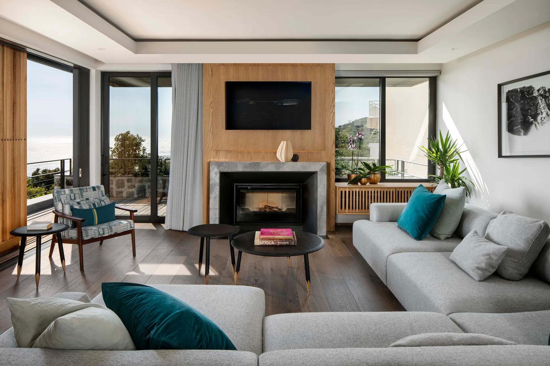 A modern living room features a gray sectional sofa, a central wood-paneled fireplace wall, and large sliding glass doors opening to an ocean view.