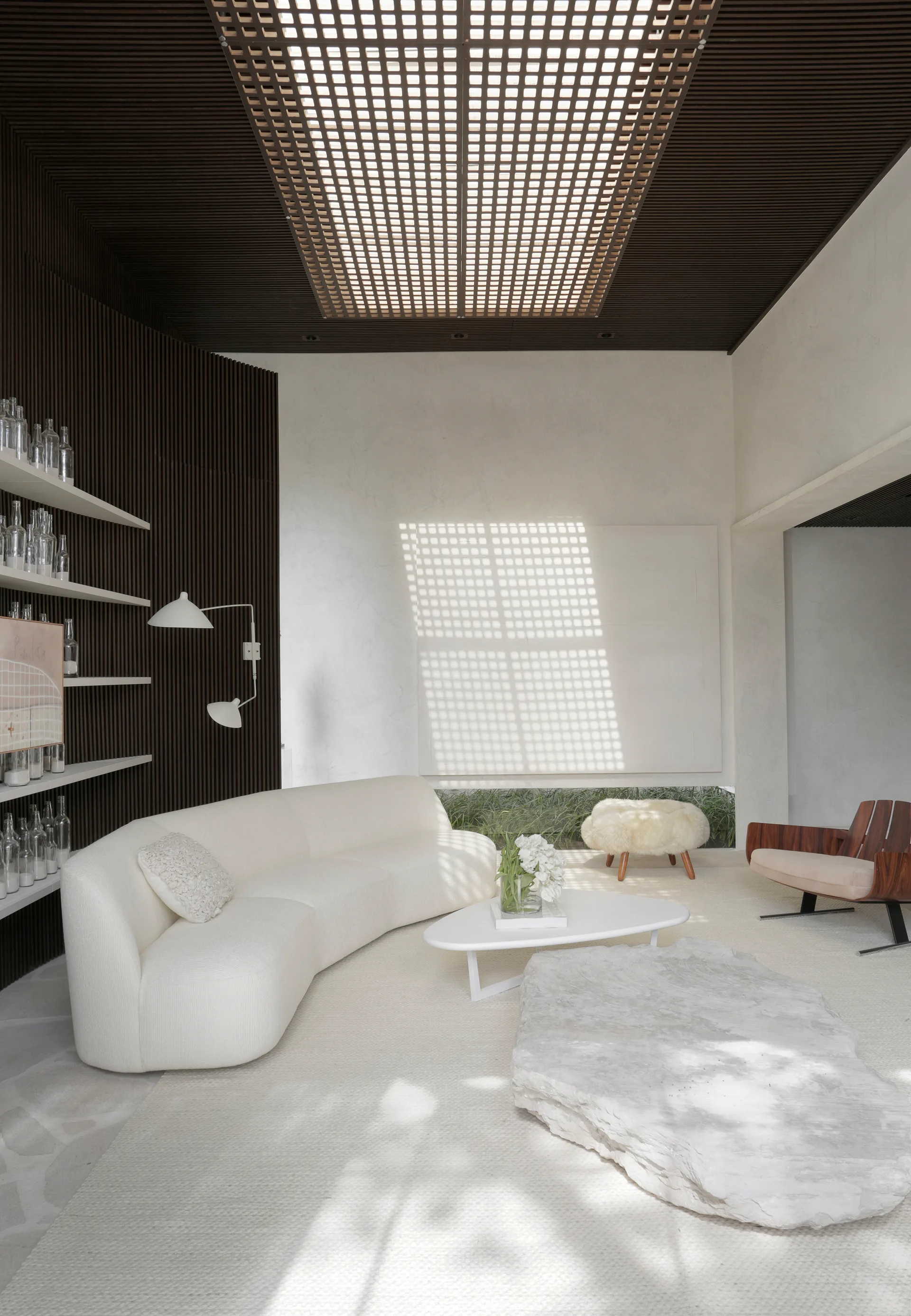 An organic minimalist living room features a curved white sofa, a dark slatted wood accent wall, and a large grid ceiling light.
