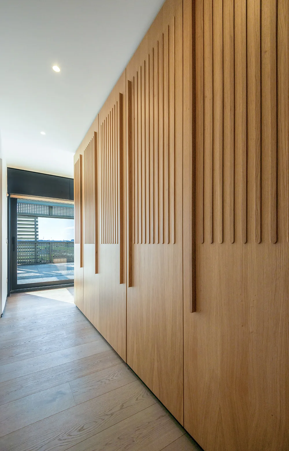 Full-height light wood built-in closets with vertical slat details line a minimalist hallway leading to a black-framed glass balcony door.