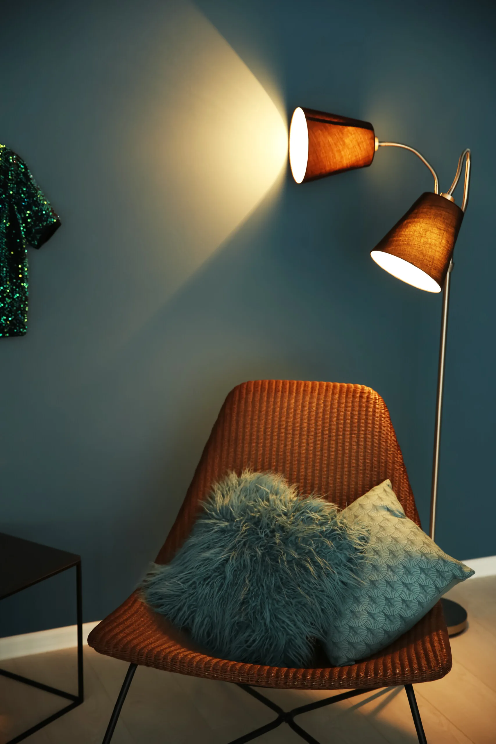 A woven wicker armchair holding two teal textured throw pillows is illuminated by a two-headed floor lamp against a dark teal wall.