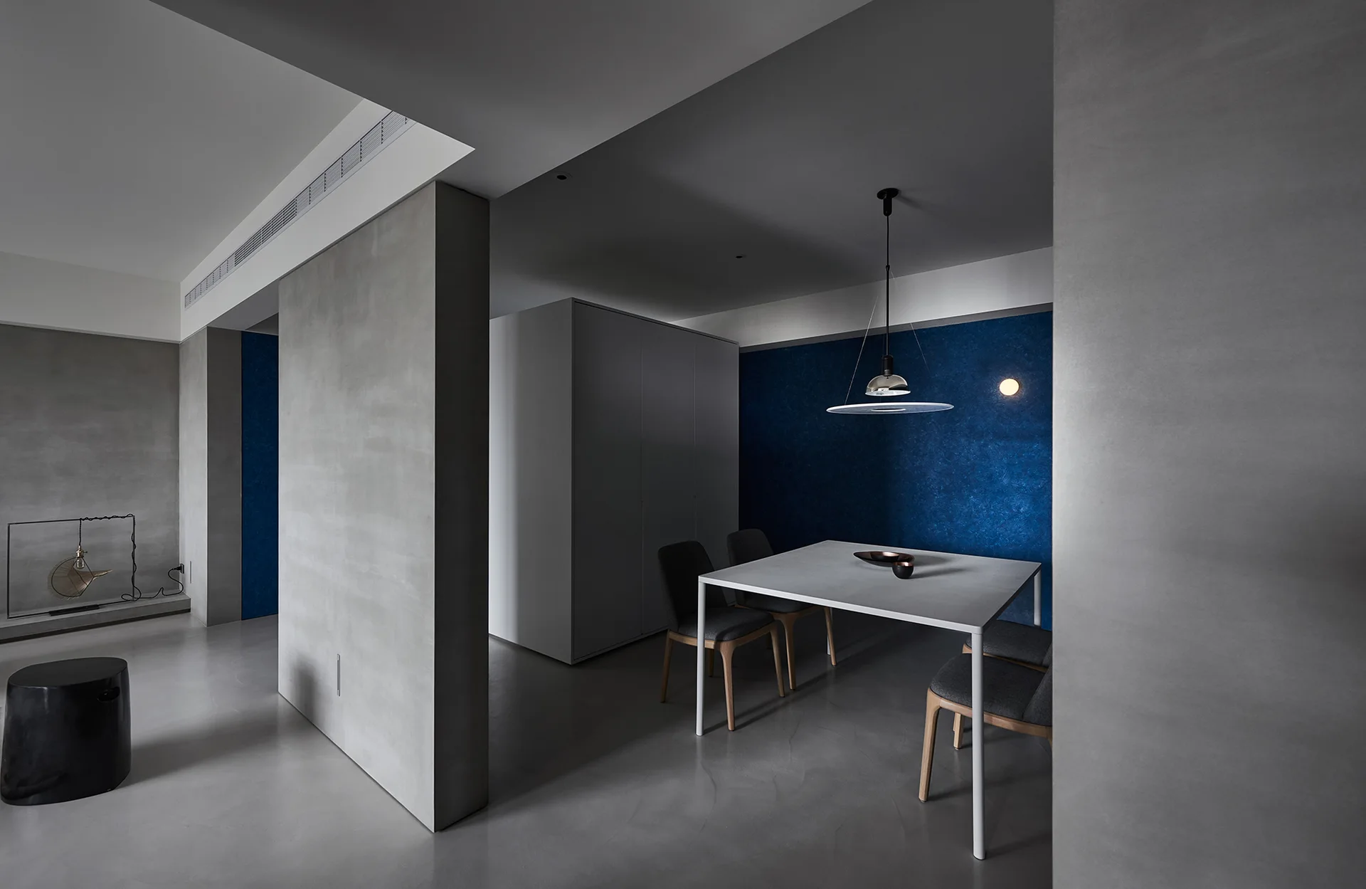 A minimalist dining area featuring a white square table, grey chairs, concrete-like walls, and a textured deep blue accent wall.