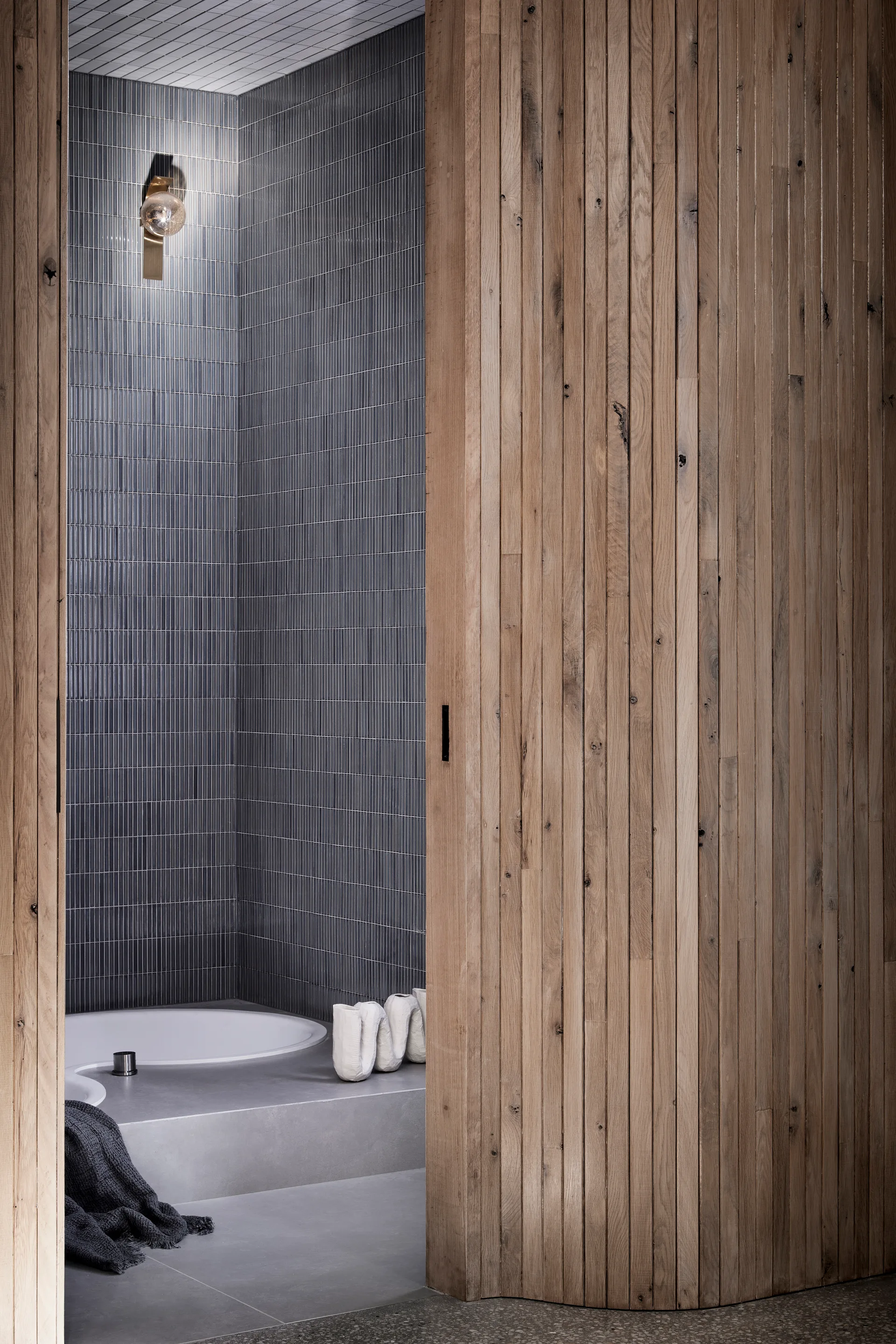 A modern bathroom with grey vertical wall tiles and a built-in white tub, partially hidden by a natural wood sliding pocket door.