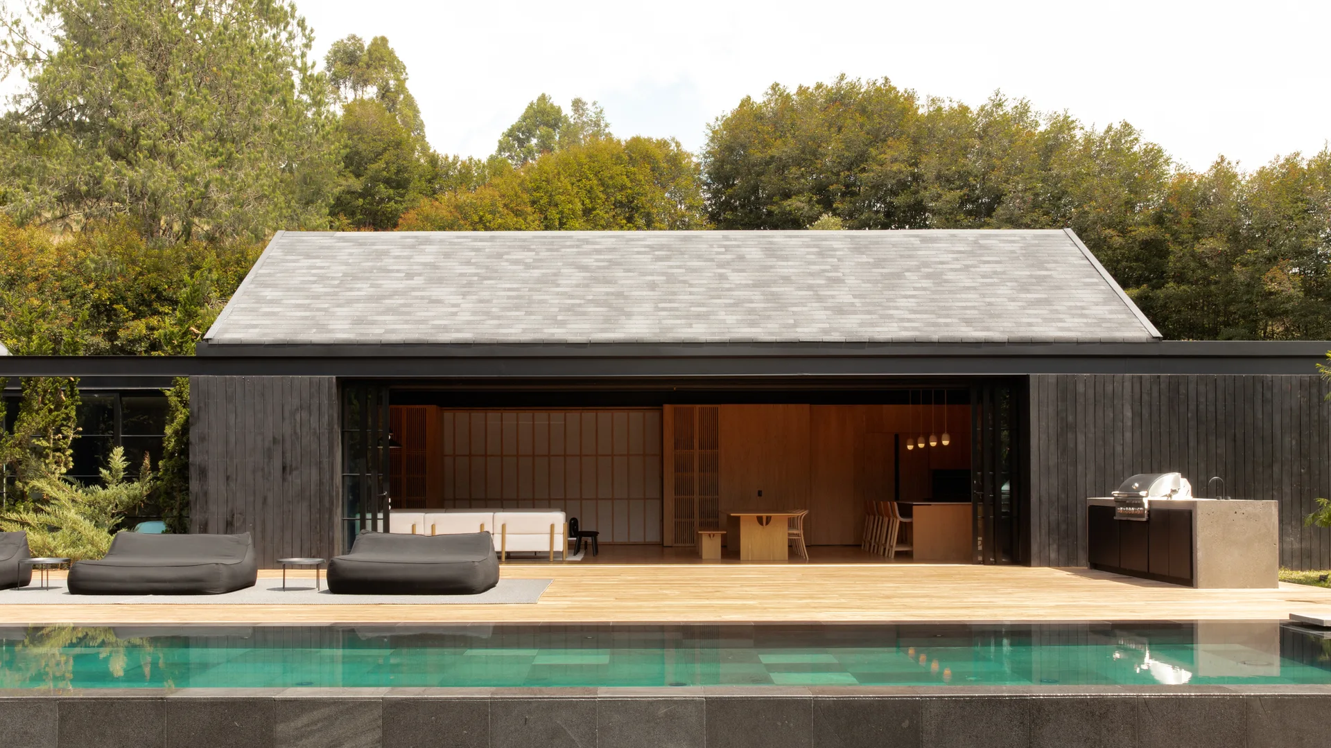 A modern pool house with dark charcoal siding stands open to a wide wooden deck, featuring a swimming pool and low-profile black lounge chairs.