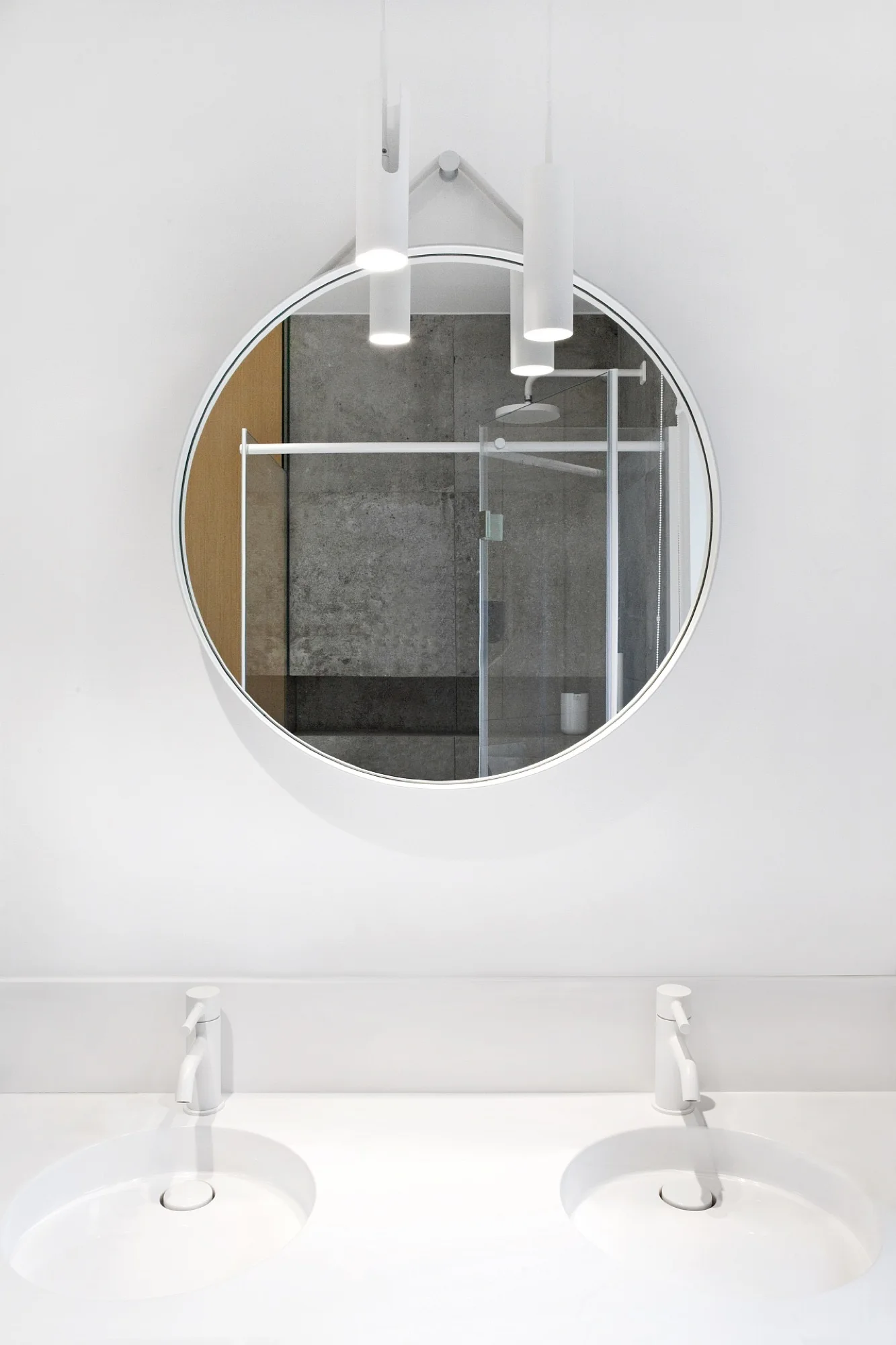 A minimalist bathroom features a white double vanity with matching white faucets beneath a large circular mirror reflecting a grey tiled shower.
