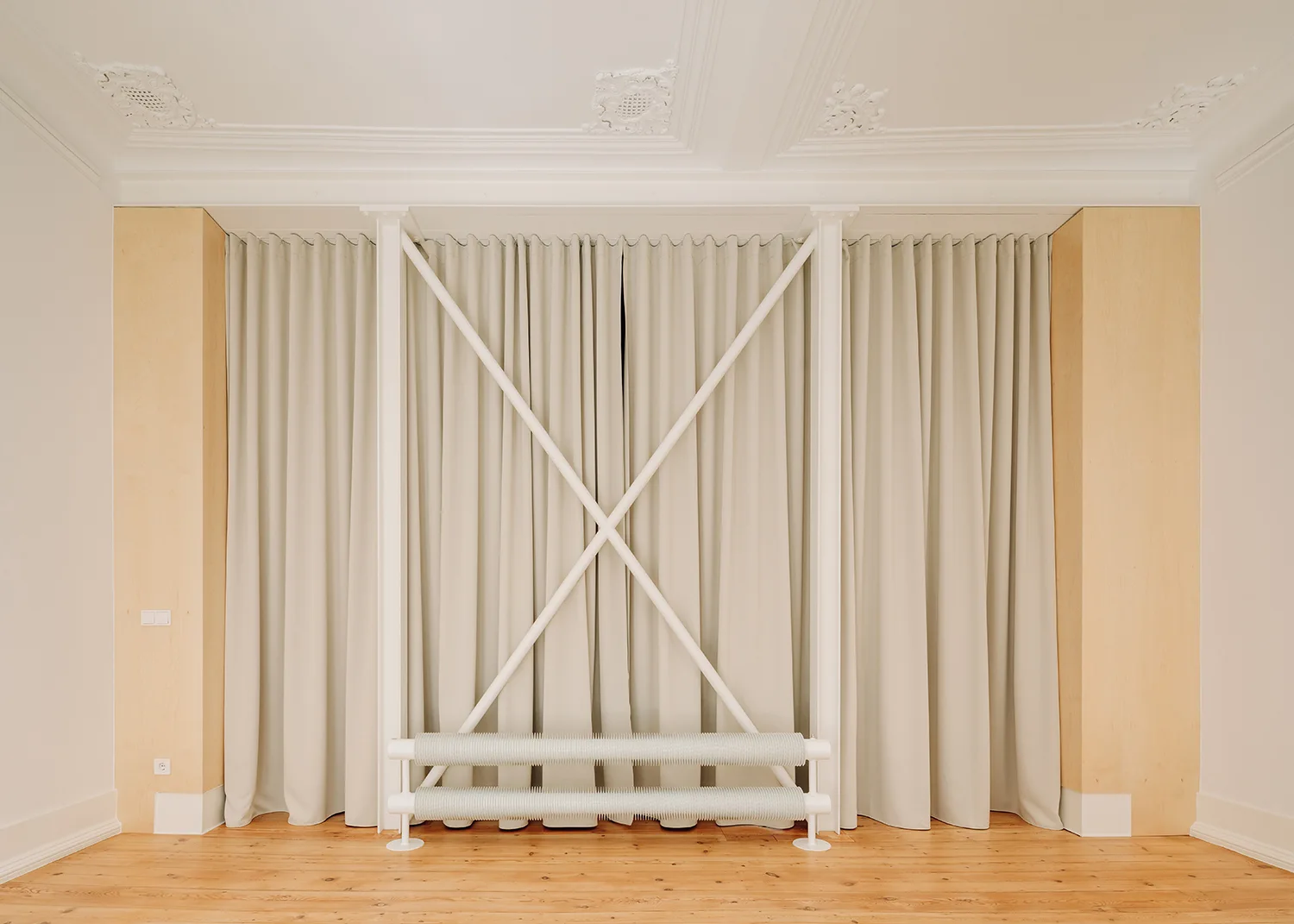 A minimalist room features light wood columns framing beige floor-to-ceiling curtains, white structural cross-bracing, and an industrial radiator.