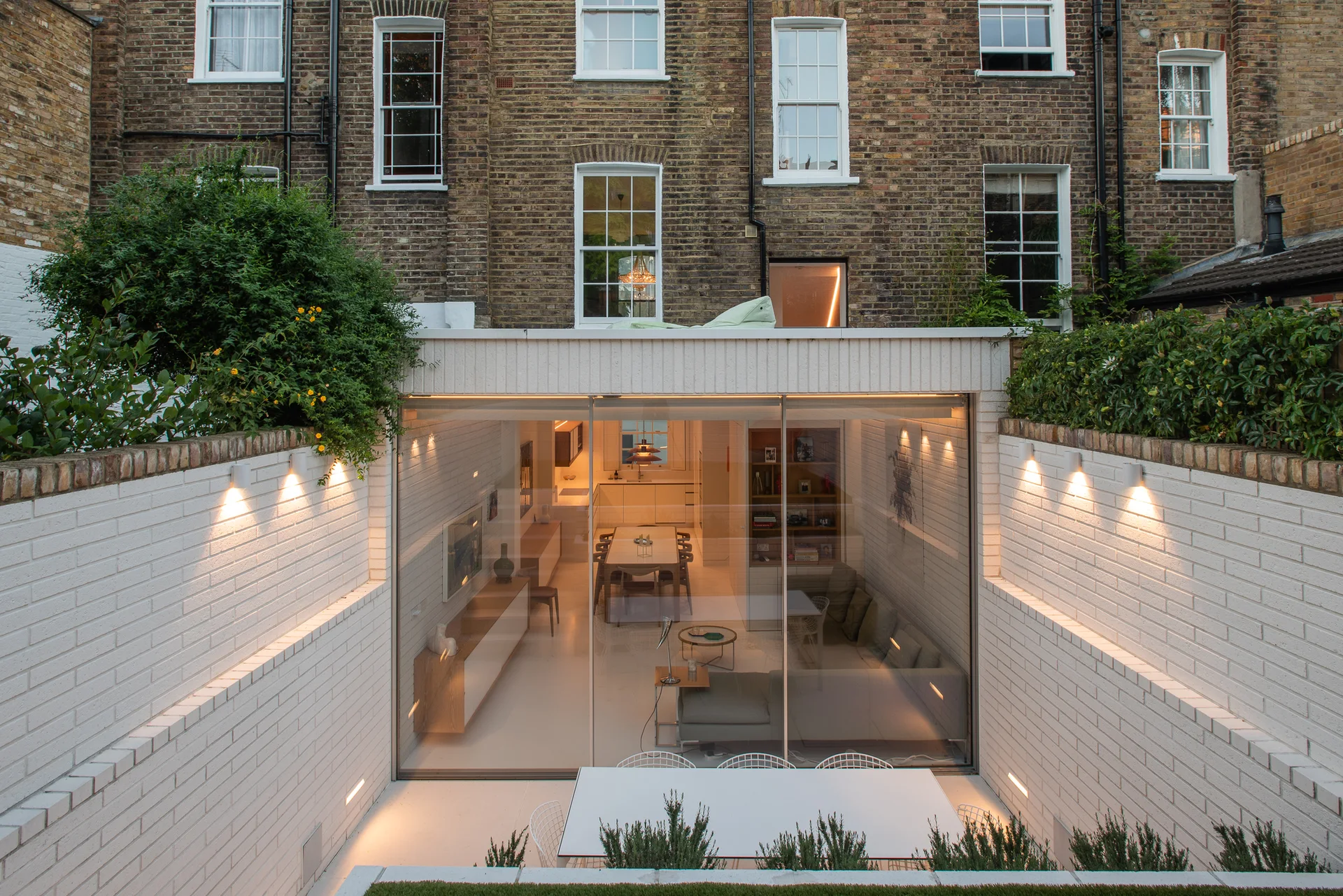 A modern glass-walled home extension opens onto a sunken patio with white-painted brick retaining walls illuminated by outdoor lights.
