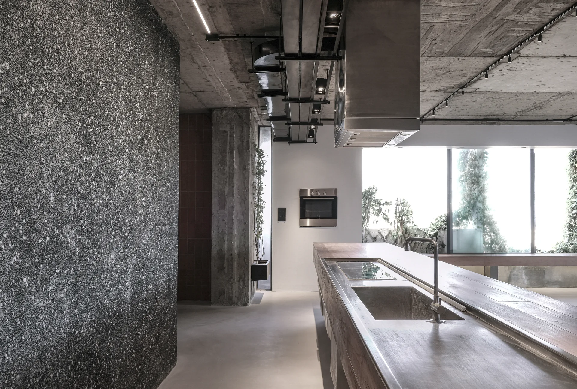 An industrial kitchen featuring a long kitchen island, an exposed concrete ceiling with utility pipes, and a dark speckled wall.