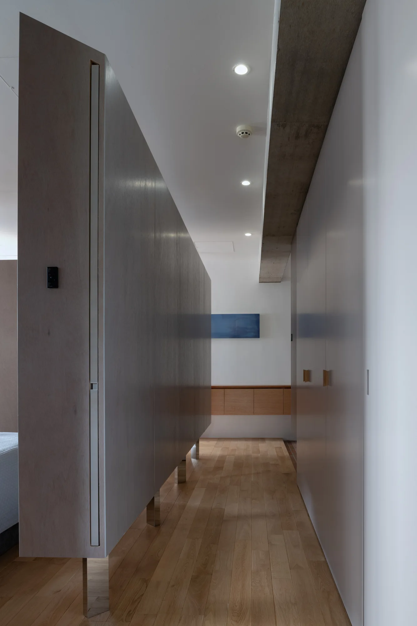A minimalist hallway with light hardwood floors, a grey wooden partition on metal legs, white closets, and an exposed concrete ceiling beam.