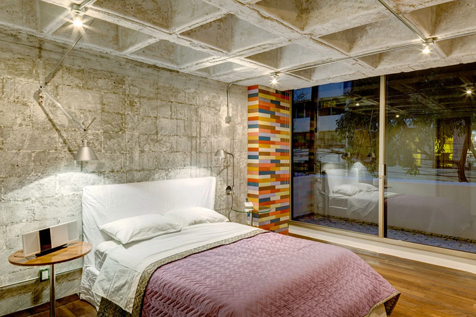 An industrial bedroom features a concrete waffle slab ceiling, raw concrete block walls, a bed with purple bedding, and a colorful tiled accent column.