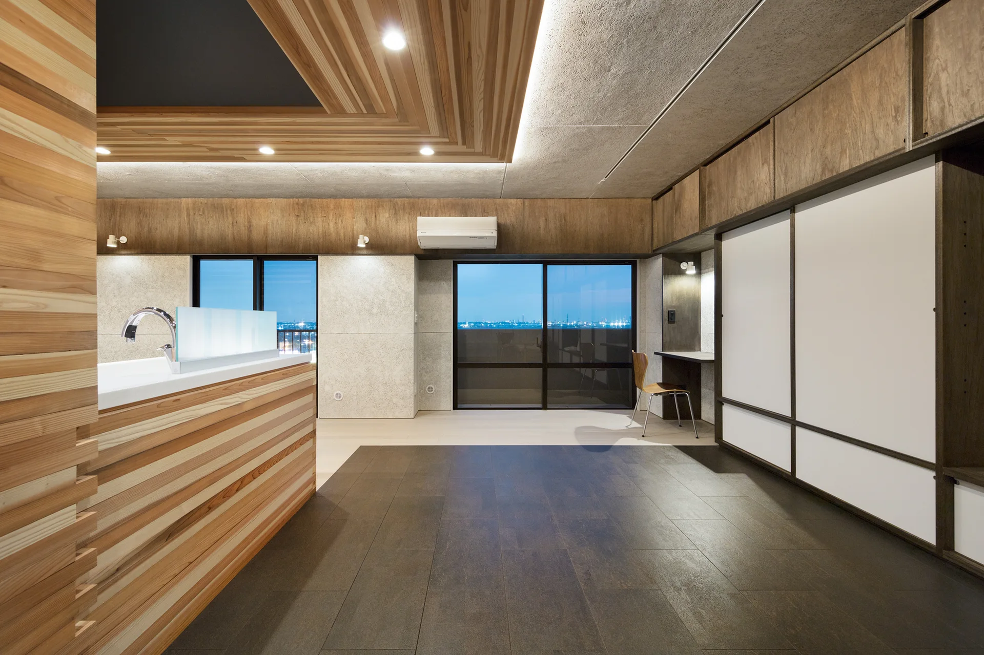 A minimalist modern room features a slatted wood ceiling, dark tiled flooring, a wooden kitchen island, and white sliding cabinet doors.