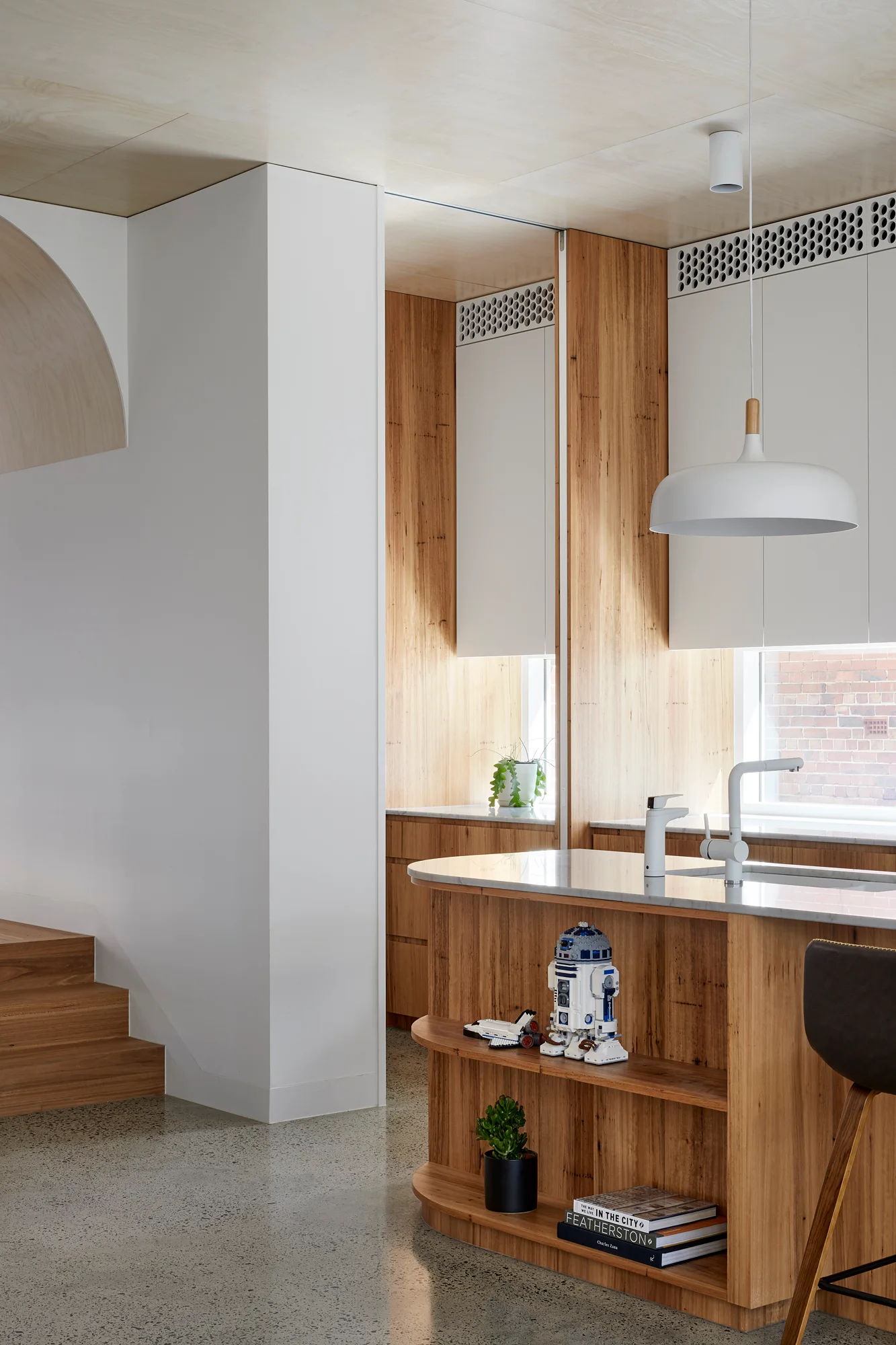 A modern kitchen features a curved wood island with open shelves, white upper cabinets, a paneled wood ceiling, and polished concrete flooring.