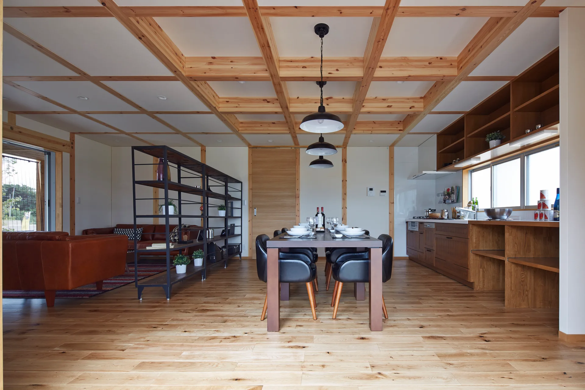 A dining table with black leather chairs sits under a wooden grid ceiling and black pendant lights in an open-concept living space.
