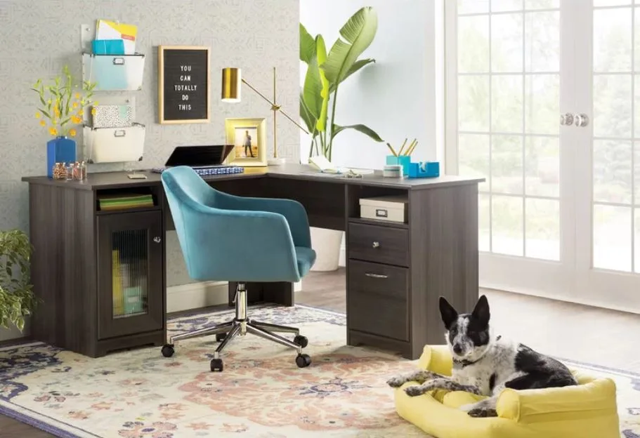 A home office features a dark wood L-shaped desk, a blue velvet swivel chair, and a patterned rug near a glass door.