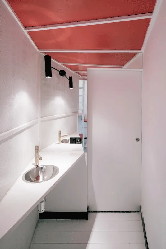A minimalist powder room features a white triangular vanity counter, a small metal sink, a wall mirror, and a bright red ceiling.