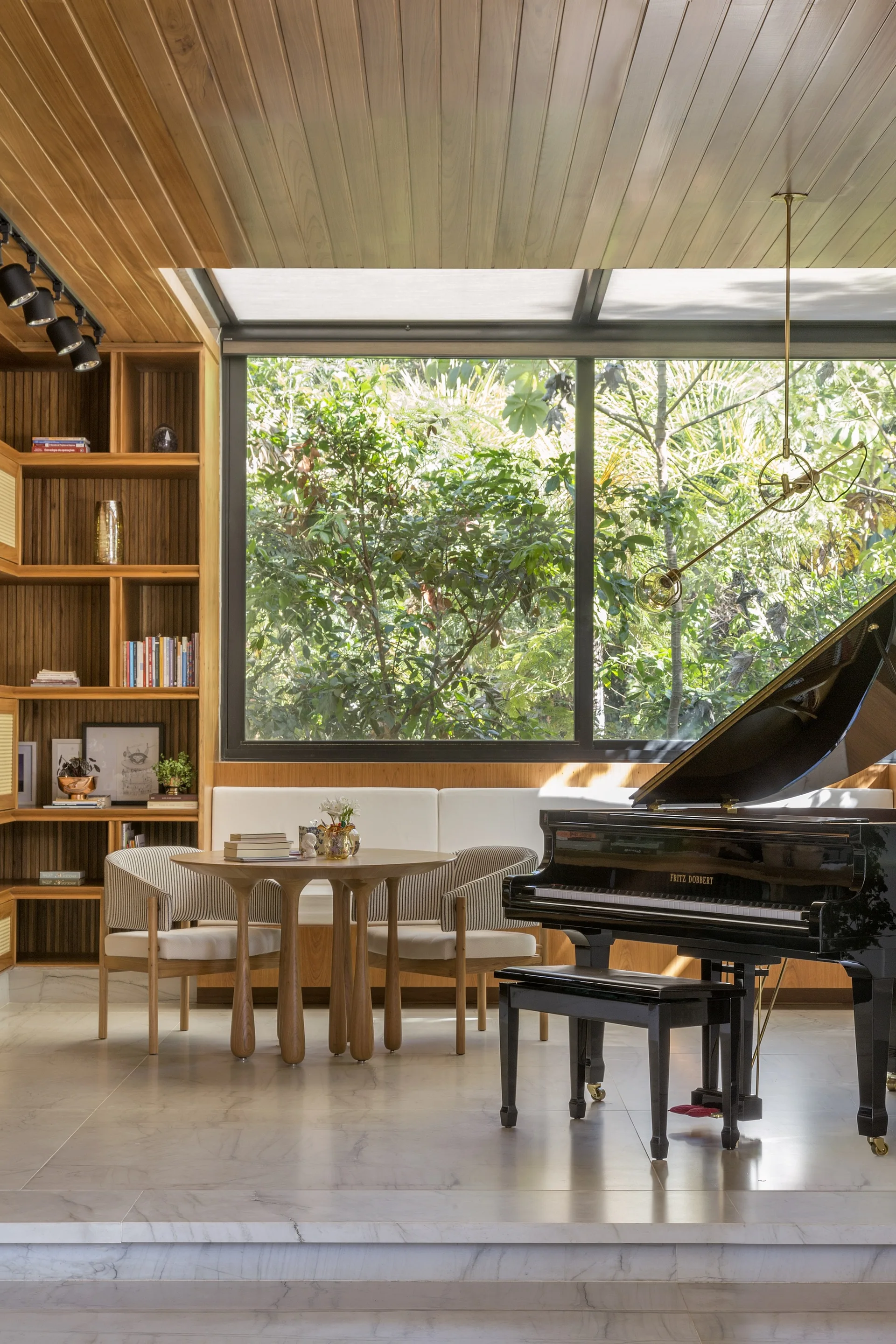 A sunlit living room features a black grand piano, a wooden dining nook, built-in shelving, and a large window looking out onto a green garden.